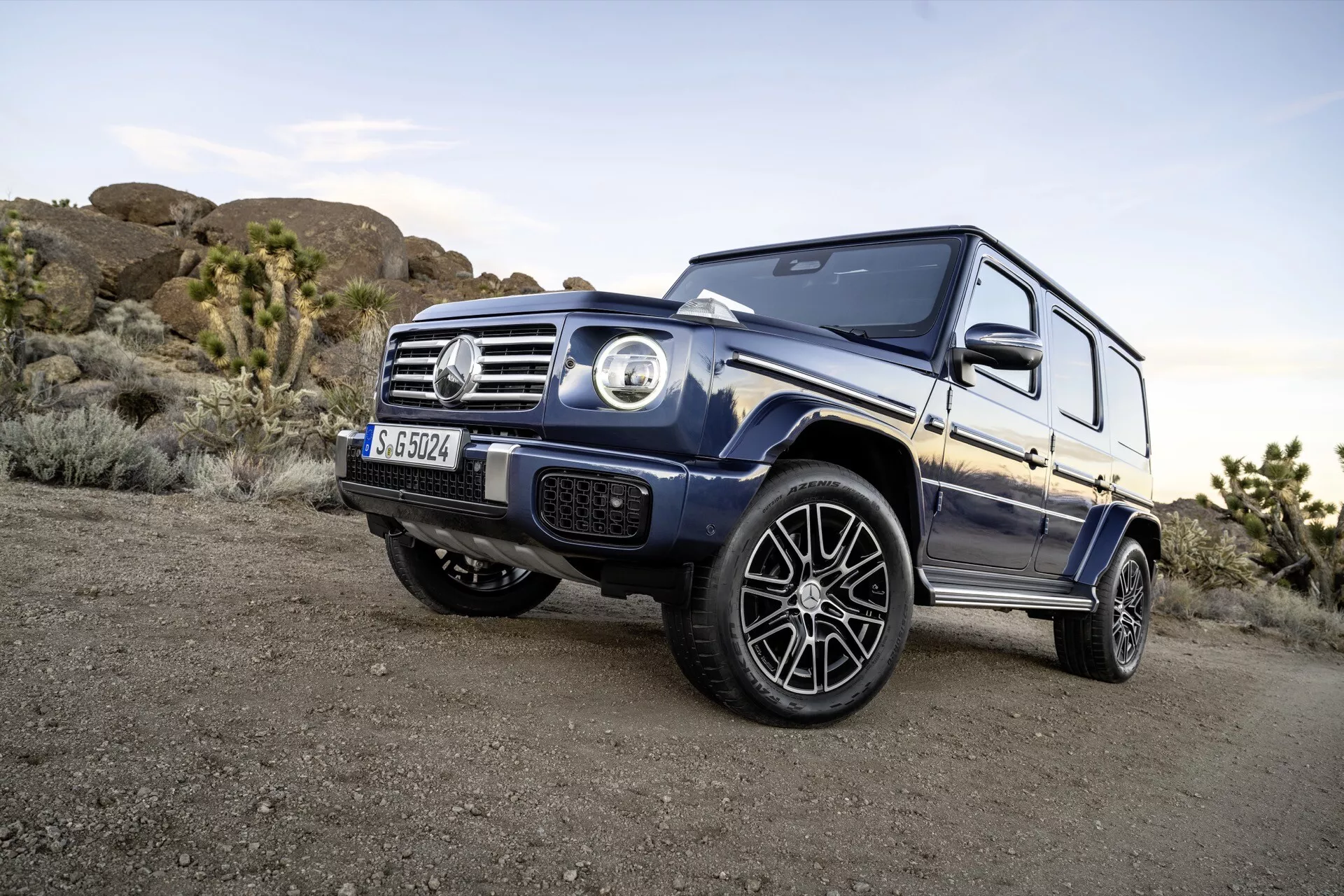 Prise de vue angulaire dynamique du SUV Mercedes-Benz Classe G 2024 soulignant sa posture puissante et ses éléments de conception détaillés tels que les phares ronds, la calandre avant avec emblème, et les arches de roue musclées contre un arrière-plan tout-terrain dans une douce lumière du soir