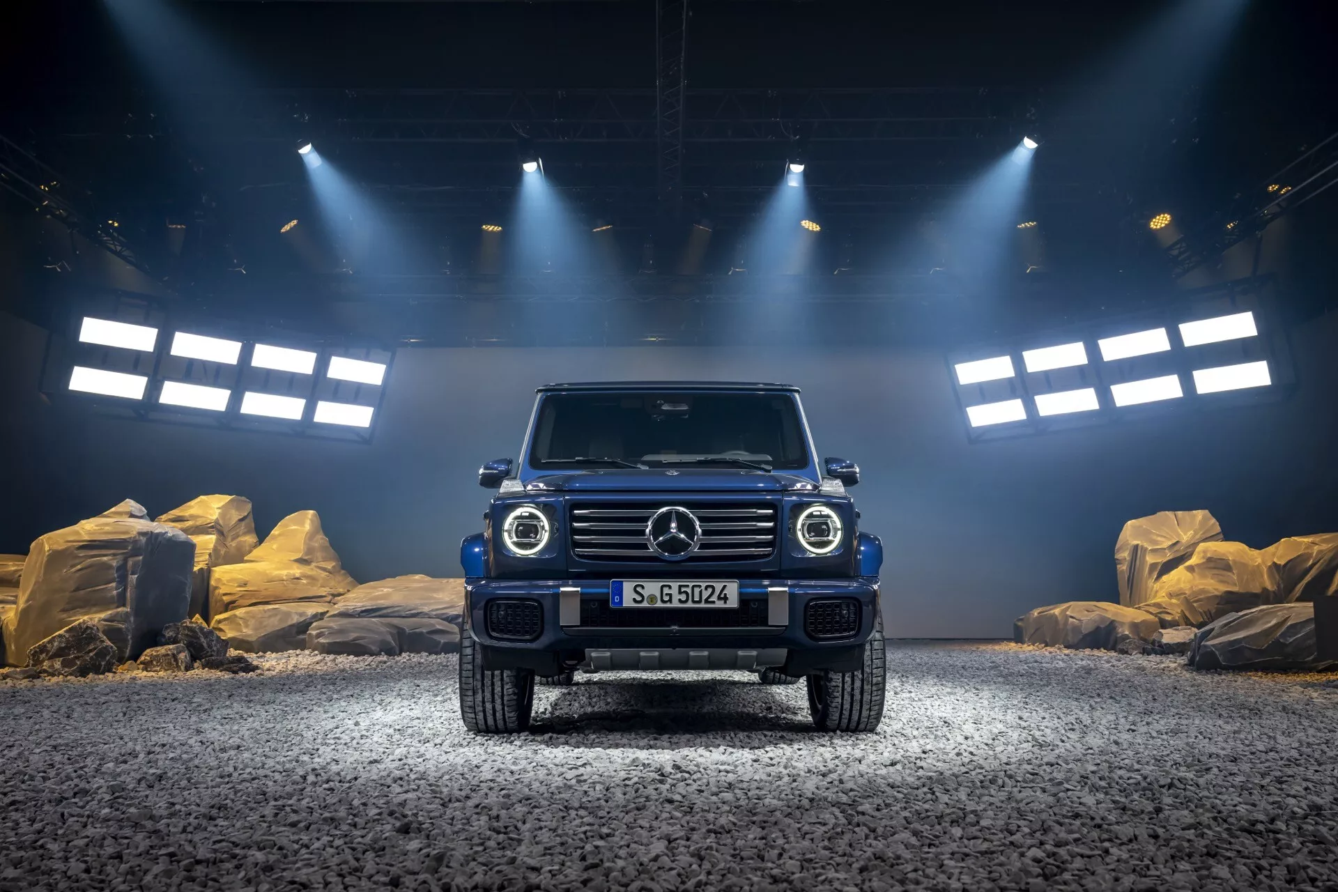 SUV de luxe tout-terrain Mercedes-Benz Classe G (G-Klasse) 2024 avec un éclairage spectaculaire mettant en valeur sa garde au sol élevée, sa grille emblématique avec emblème, ses phares ronds à LED, ses feux de jour rectangulaires, son pare-chocs avant robuste et son design musclé du capot sur une surface en gravier avec un fond de studio d'aventure