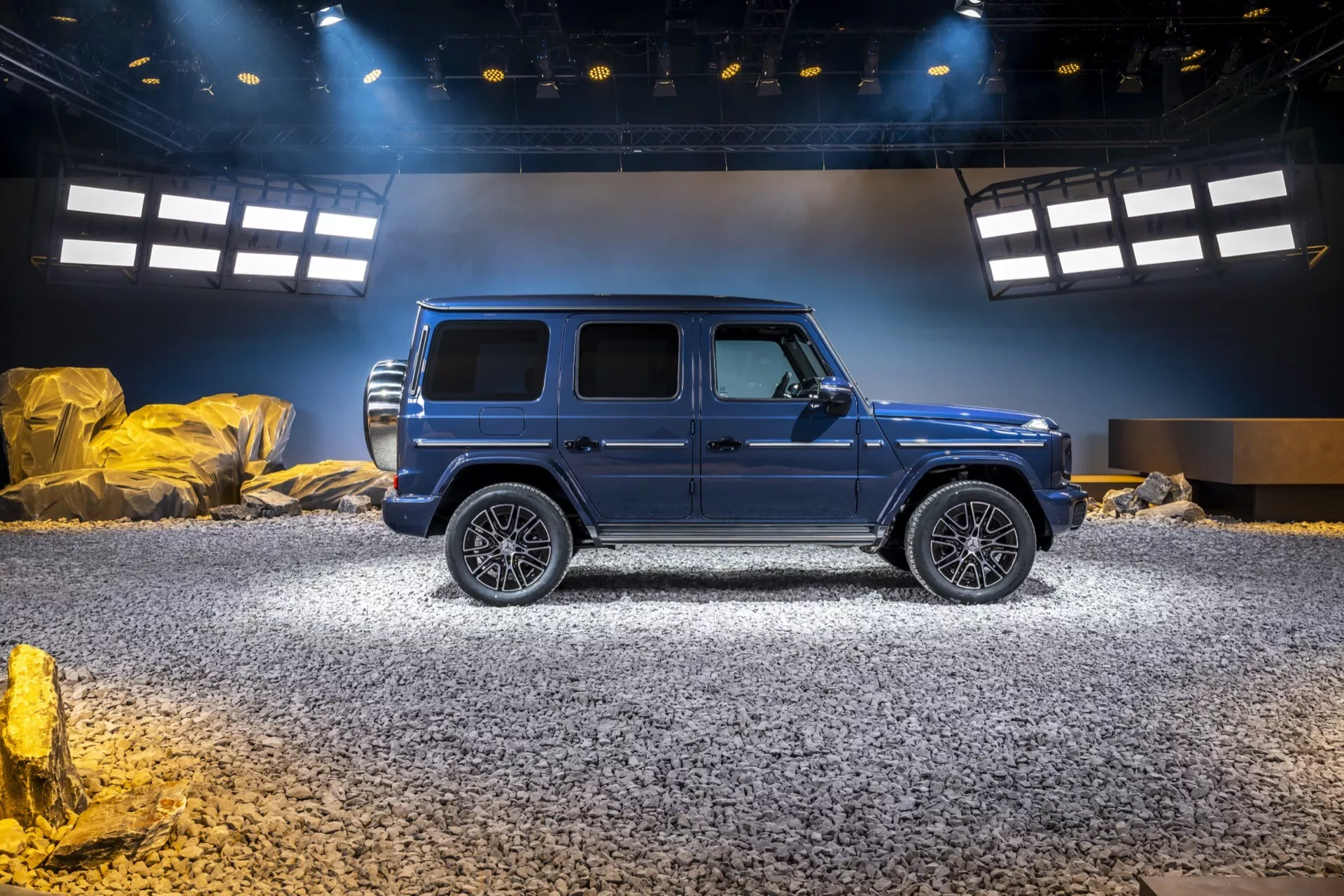 SUV de luxe Mercedes-Benz Classe G 2024 en bleu métallique démontrant des capacités tout-terrain avec de grandes roues en alliage, des marches latérales et un toit ouvrant, centré dans un environnement d'exposition tout-terrain simulé