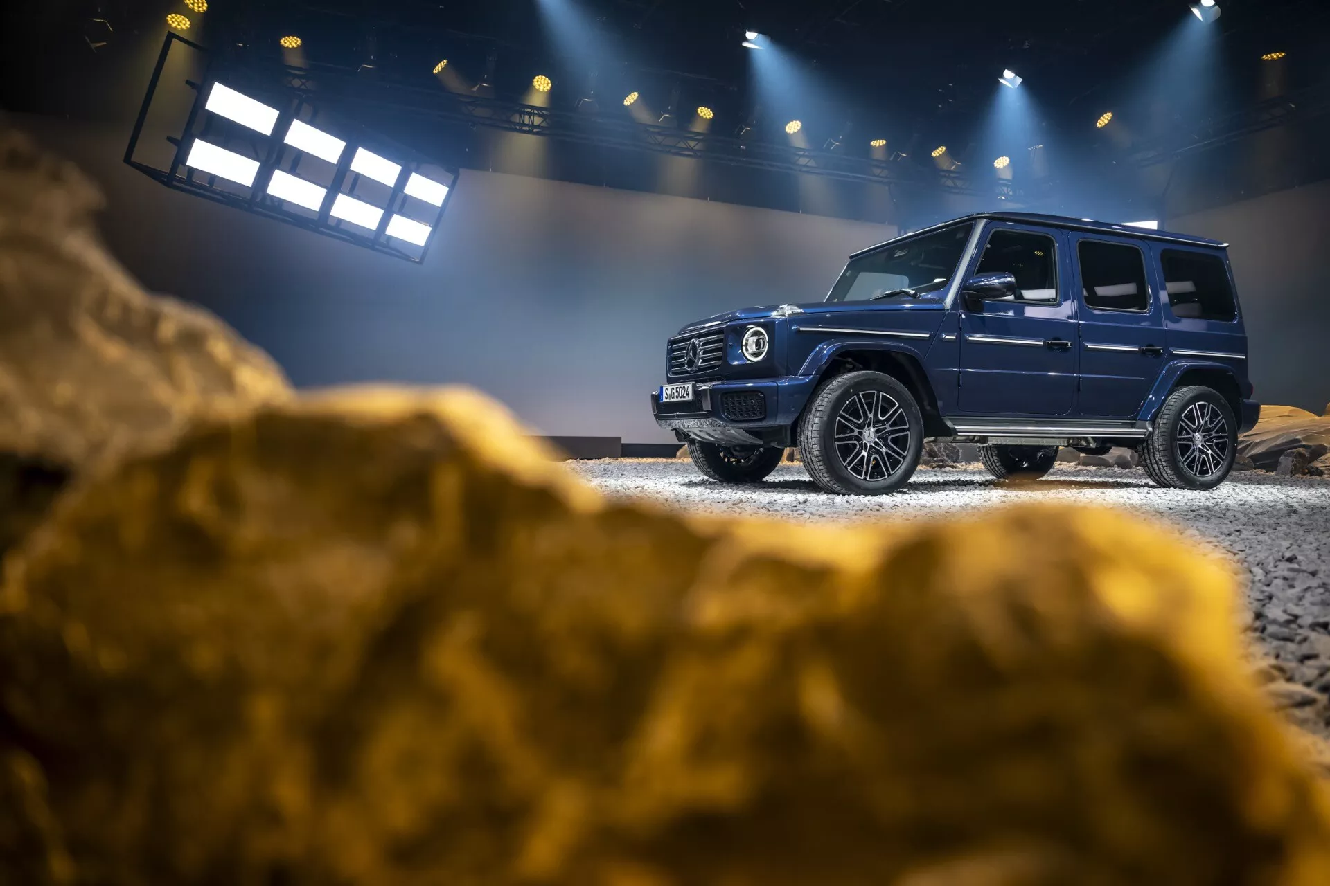 SUV de luxe Mercedes-Benz Classe G 2024 présenté en intérieur sur un terrain tout-terrain simulé, mettant en avant le profil latéral, la grille avant et les caractéristiques technologiques avancées avec un éclairage contrôlé et un fond robuste
