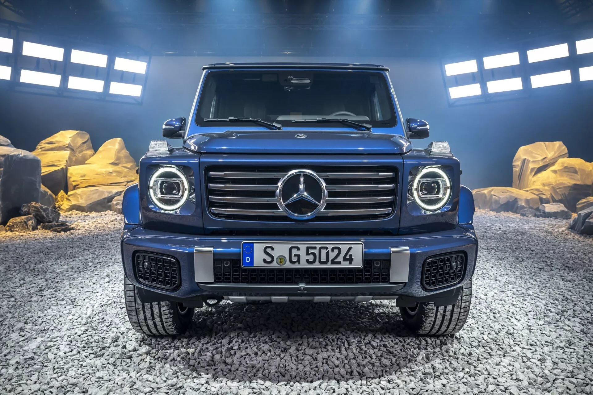 Vue de face de la Mercedes-Benz Classe G 2024 montrant la grille iconique, les phares LED, la conception robuste du capot, et le pare-chocs avant prêt pour le tout-terrain avec plaque de protection, capturée dans un cadre naturel en plein air