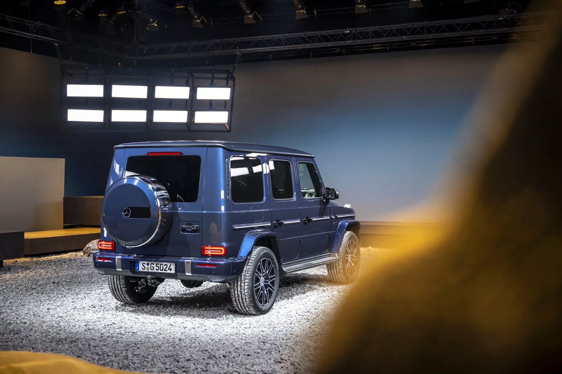 Vue latérale et partielle arrière du SUV de luxe Mercedes-Benz Classe G 2024, soulignant les caractéristiques de conception tout-terrain, les roues en alliage et les vitres teintées dans un cadre d'exposition intérieure