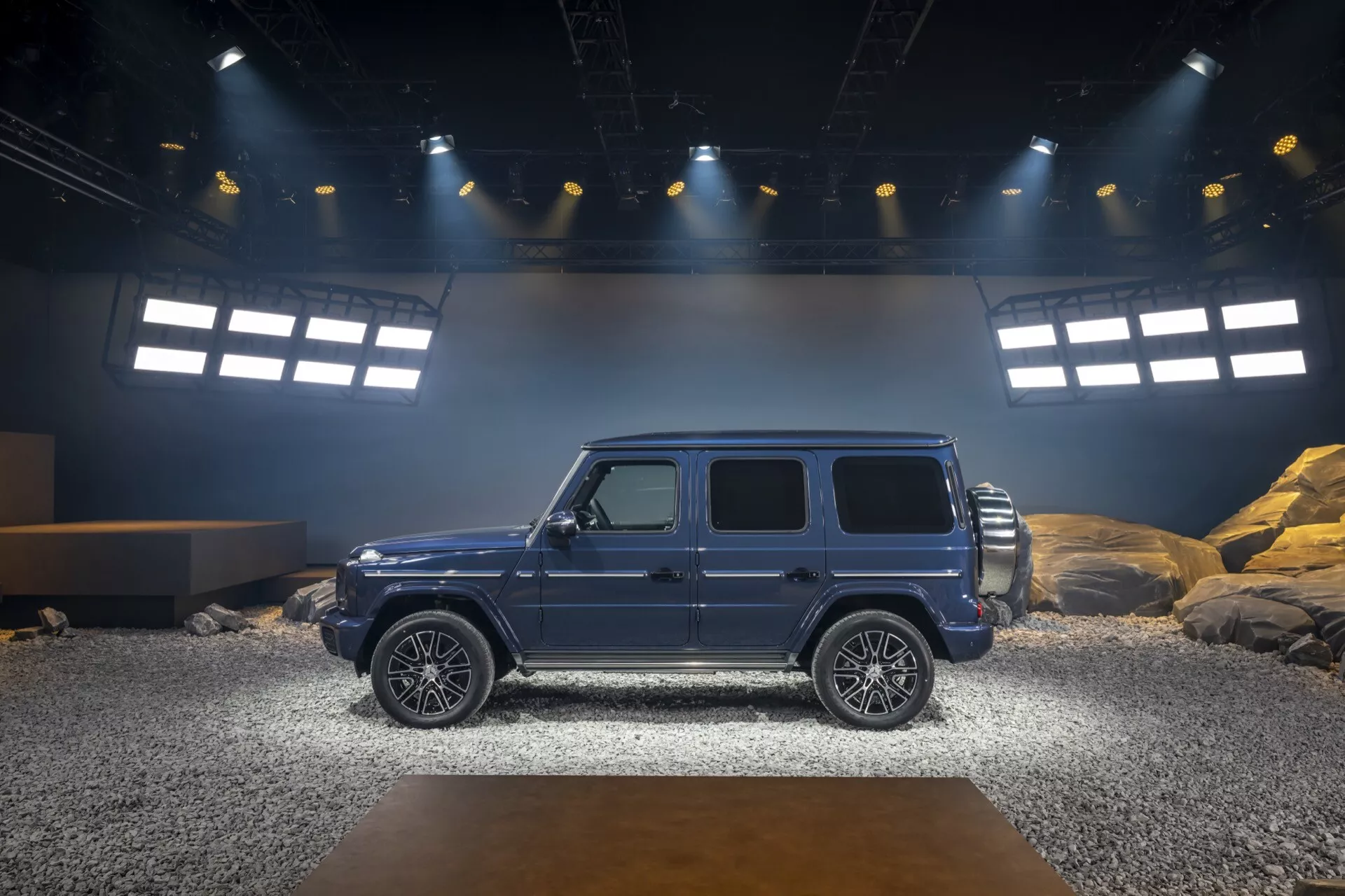Profil latéral du SUV de luxe Mercedes-Benz G-Klasse 465 2024 sur une scène intérieure simulant un terrain tout-terrain, mettant en avant l'esthétique du design, la stabilité et les caractéristiques technologiques avancées sous un éclairage vif de projecteurs