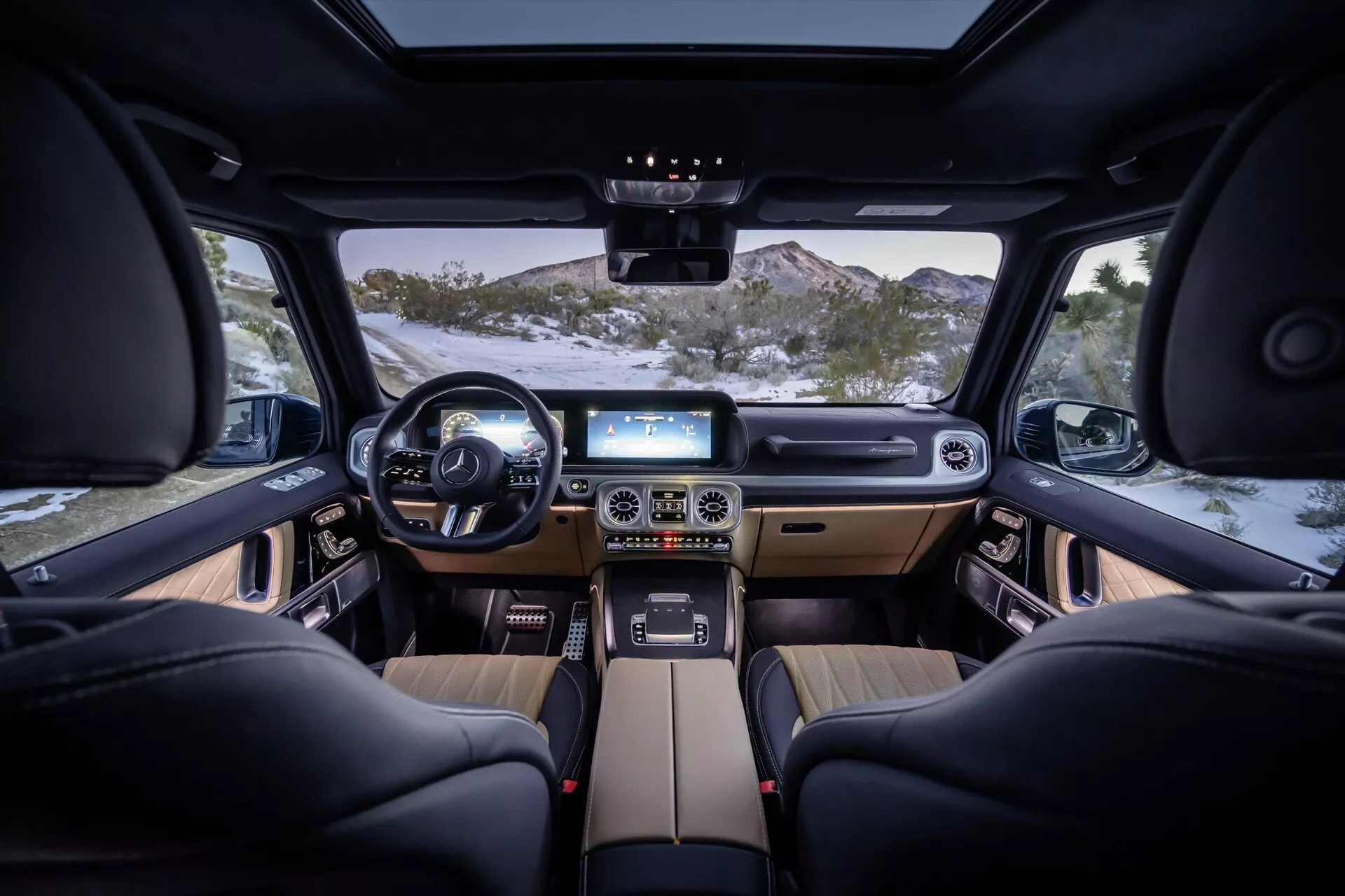 Vue intérieure du Mercedes-Benz Classe G 2024 depuis l'arrière-centre, présentant un volant en cuir multifonction, deux affichages numériques sur le tableau de bord, la console centrale avec pavé tactile, le levier de vitesse automatique, des sièges en cuir noir à réglage électronique, des accents en bois, un éclairage d'ambiance et une vue claire à travers le pare-brise avant