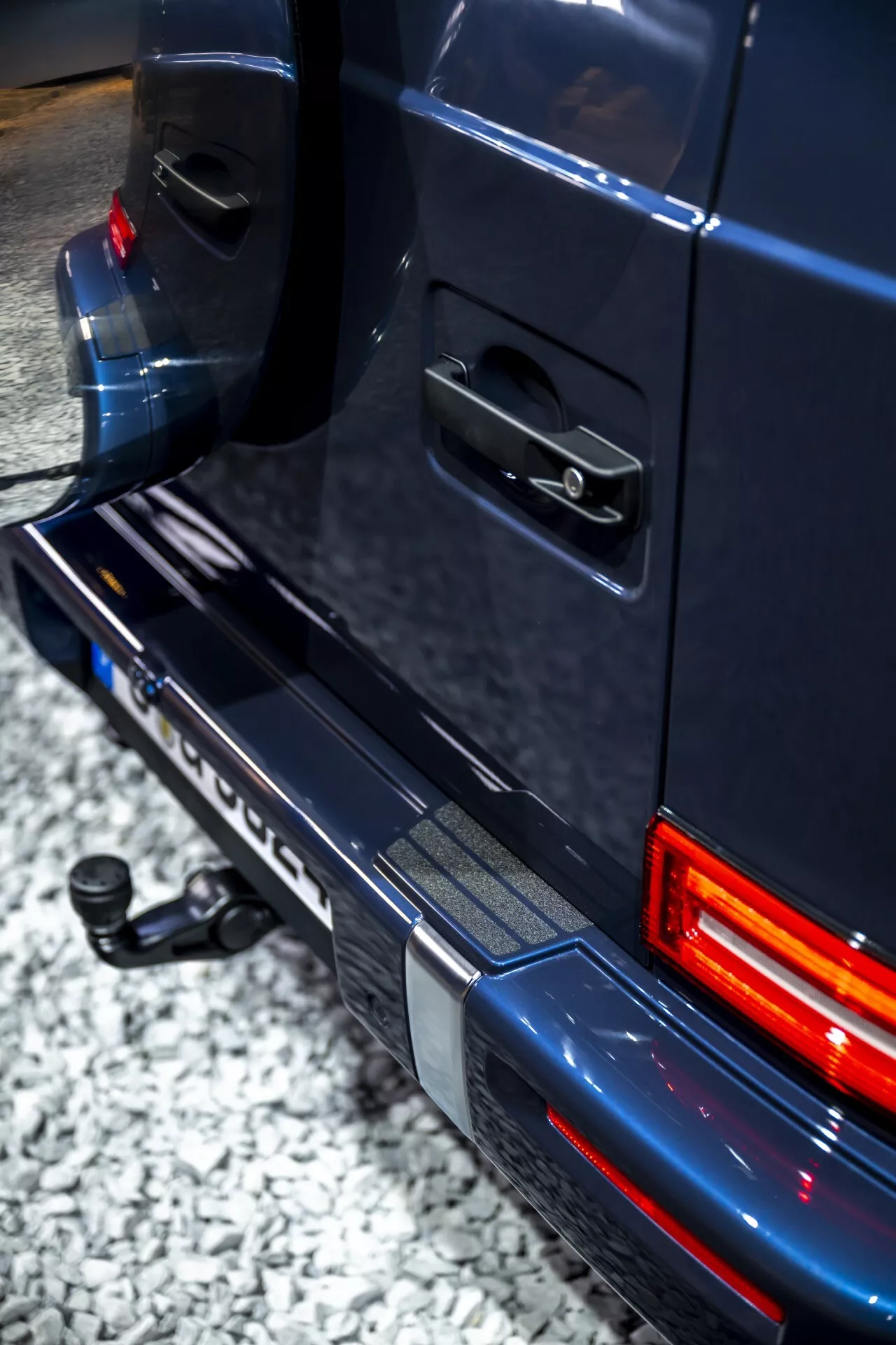 Vue en angle rapprochée mettant en lumière la section arrière d'une Mercedes-Benz G-Klasse 2024, montrant le côté droit du feu arrière, du pare-chocs arrière, et des détails de la porte avec éclairage LED et caractéristiques pour le remorquage.