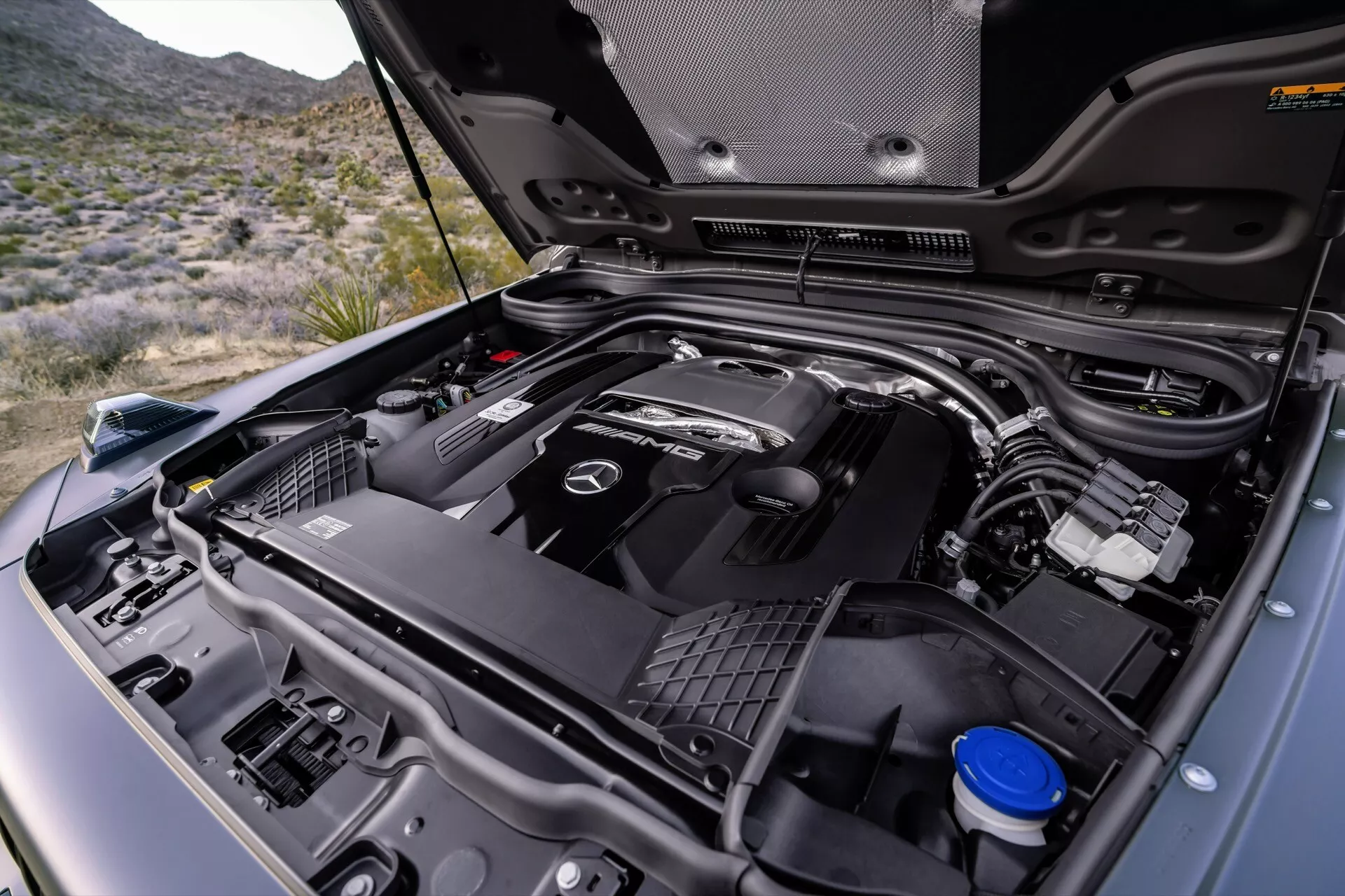 Vue rapprochée du compartiment moteur de la Mercedes-Benz G-Klasse 2024, montrant l’emblème de la marque sur le couvercle du moteur, vue détaillée de diverses parties du moteur, dont l'admission d'air et le réservoir de liquide de refroidissement, sur fond extérieur