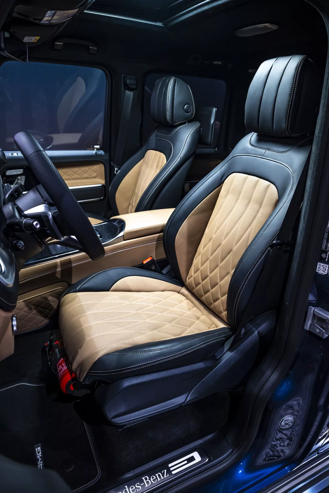 Vue intérieure luxueuse de la Mercedes-Benz G-Klasse 2024 présentant un siège conducteur détaillé avec cuir matelassé tan et noir, commandes de réglage électrique avec fonction mémoire, seuil de porte illuminé avec logo, matériaux de haute qualité et éléments de technologie avancée