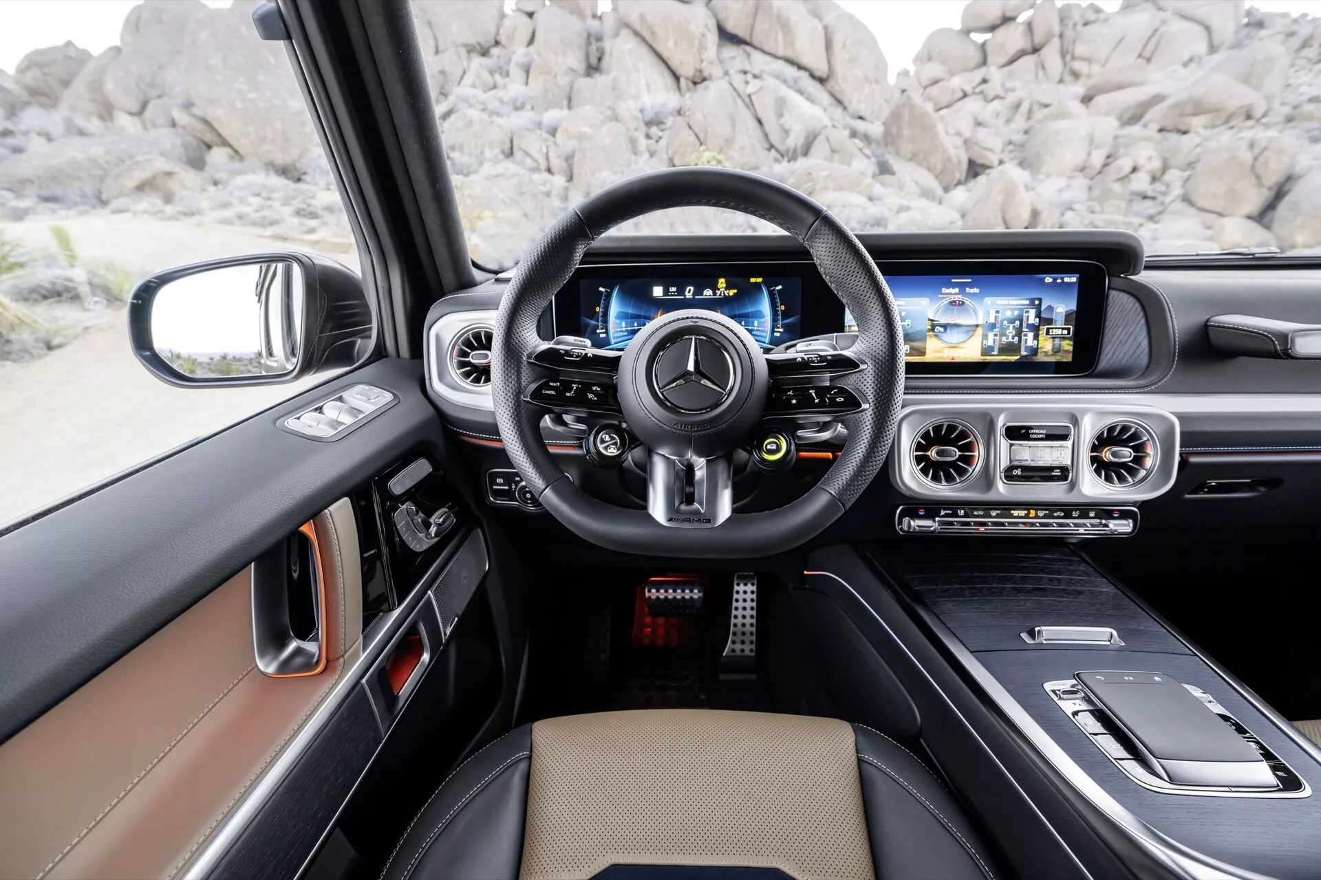 Vue intérieure depuis le siège du conducteur de la Mercedes-Benz Classe G (G-Klasse) 2024 montrant un volant multifonction en cuir, un tableau de bord numérique, un large écran d'infodivertissement avec navigation, et un design intérieur de luxe en tan et noir avec des accents de métal brossé et éventuellement des inserts en bois