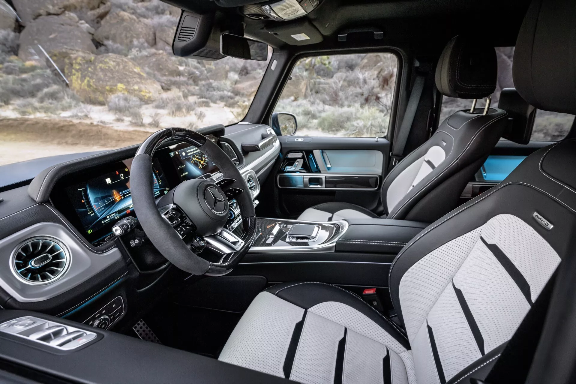 Vue intérieure de la Mercedes-Benz Classe G (G-Klasse) 2024 depuis le côté passager avant mettant en lumière l'espace spacieux de la cabine avec angle large, volant multifonction moderne, tableau de bord numérique, écran tactile d'infodivertissement avec navigation GPS, système de contrôle climatique automatique, bouches d'air style turbine, sellerie en cuir haut de gamme avec coutures contrastées, et sièges avant sportifs avec renforts latéraux
