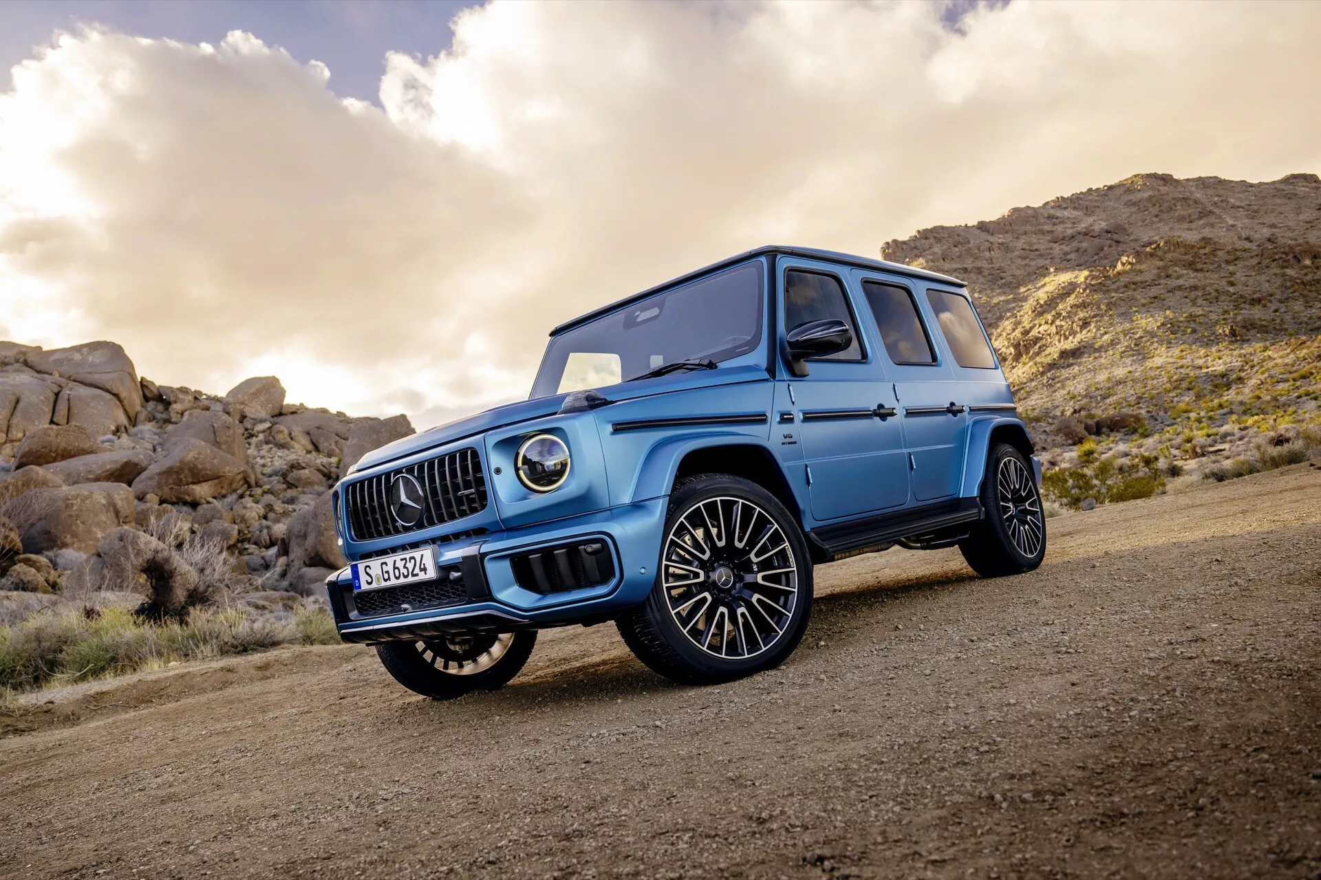 SUV de luxe Mercedes-Benz Classe G 2024 démontrant son élégance robuste et ses capacités tout-terrain sur un chemin de terre, présentant une grille avant distinctive avec un emblème central, des roues en alliage à multiples rayons pour un mélange de performance et de luxe, une peinture brillante avec un langage de design angulaire, des phares LED modernes, le tout sur un fond de terrain rocheux sous un ciel partiellement nuageux