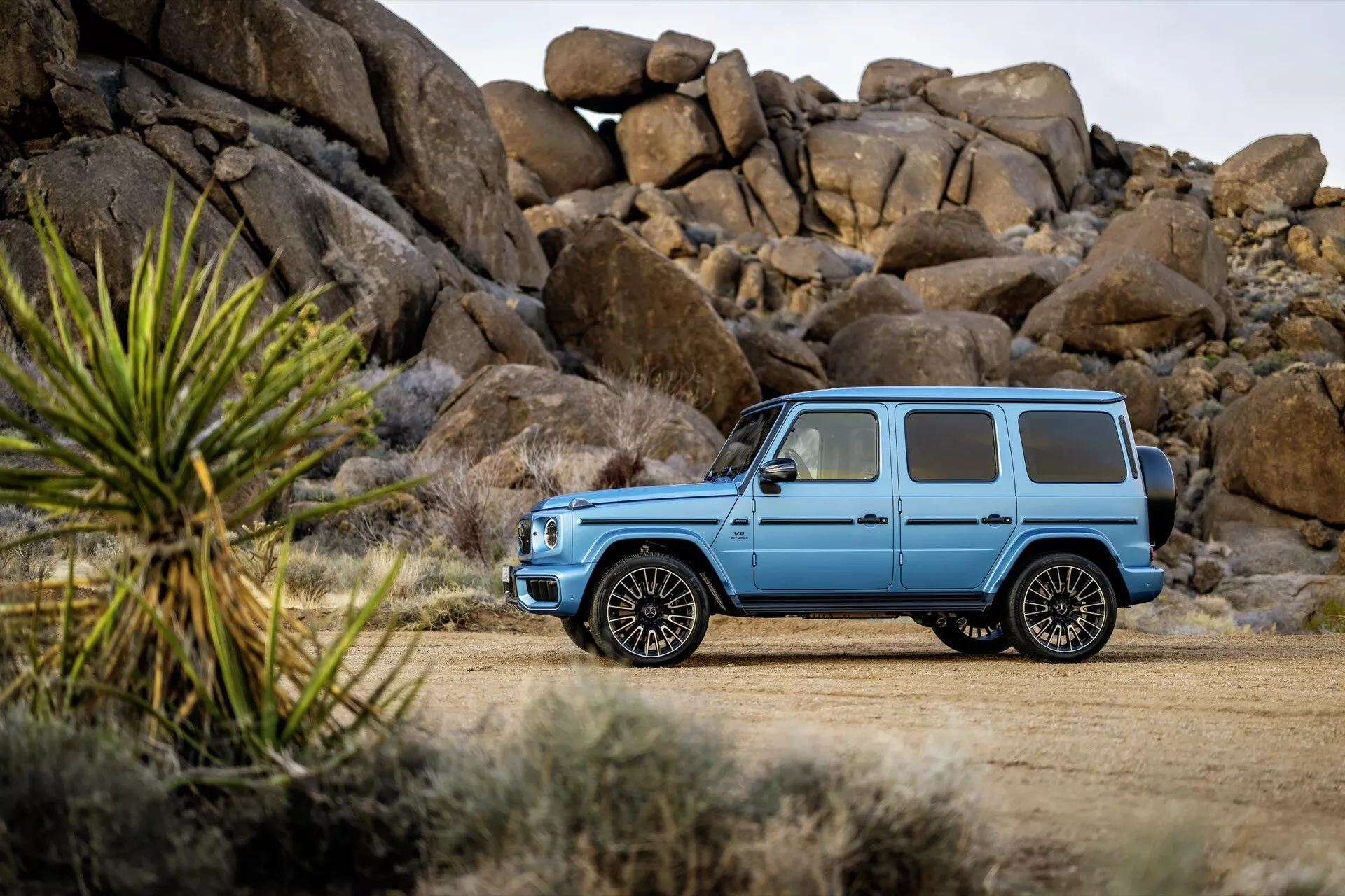 SUV de luxe Mercedes-Benz Classe G (G 465) 2024 en bleu mat clair, montrant son design robuste et sa grande garde au sol sur un chemin de terre, avec des roues en alliage complexes et des vitres teintées pour la vie privée devant un paysage accidenté.