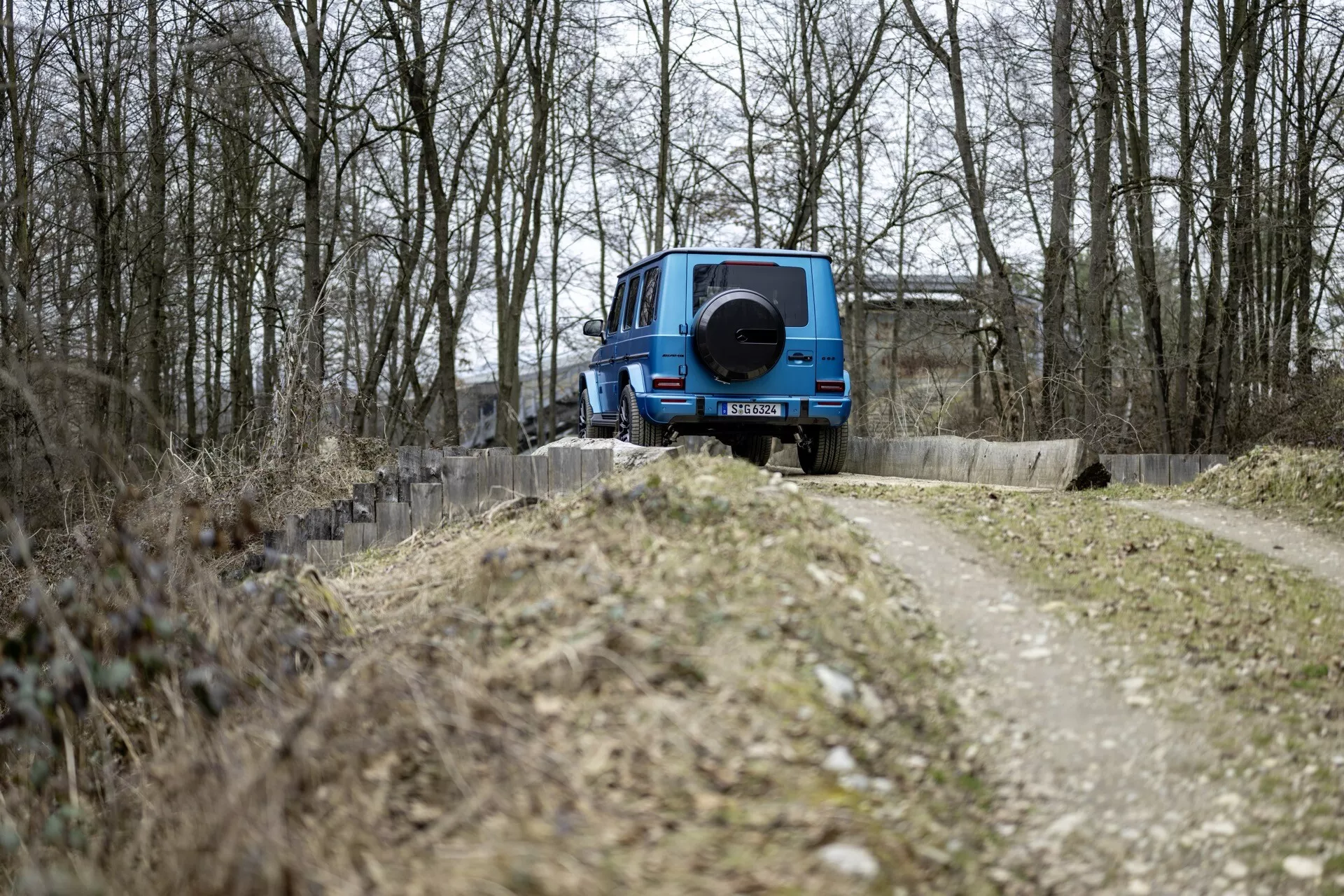 SUV Mercedes-Benz Classe G 2024 en bleu, positionné avec l'arrière face à la caméra à côté d'un chemin de terre étroit parmi des arbres sans feuilles, affichant une conception tout-terrain avec roue de secours montée à l'arrière, sous un éclairage couvert sans caractéristiques technologiques visibles sur l'extérieur arrière.