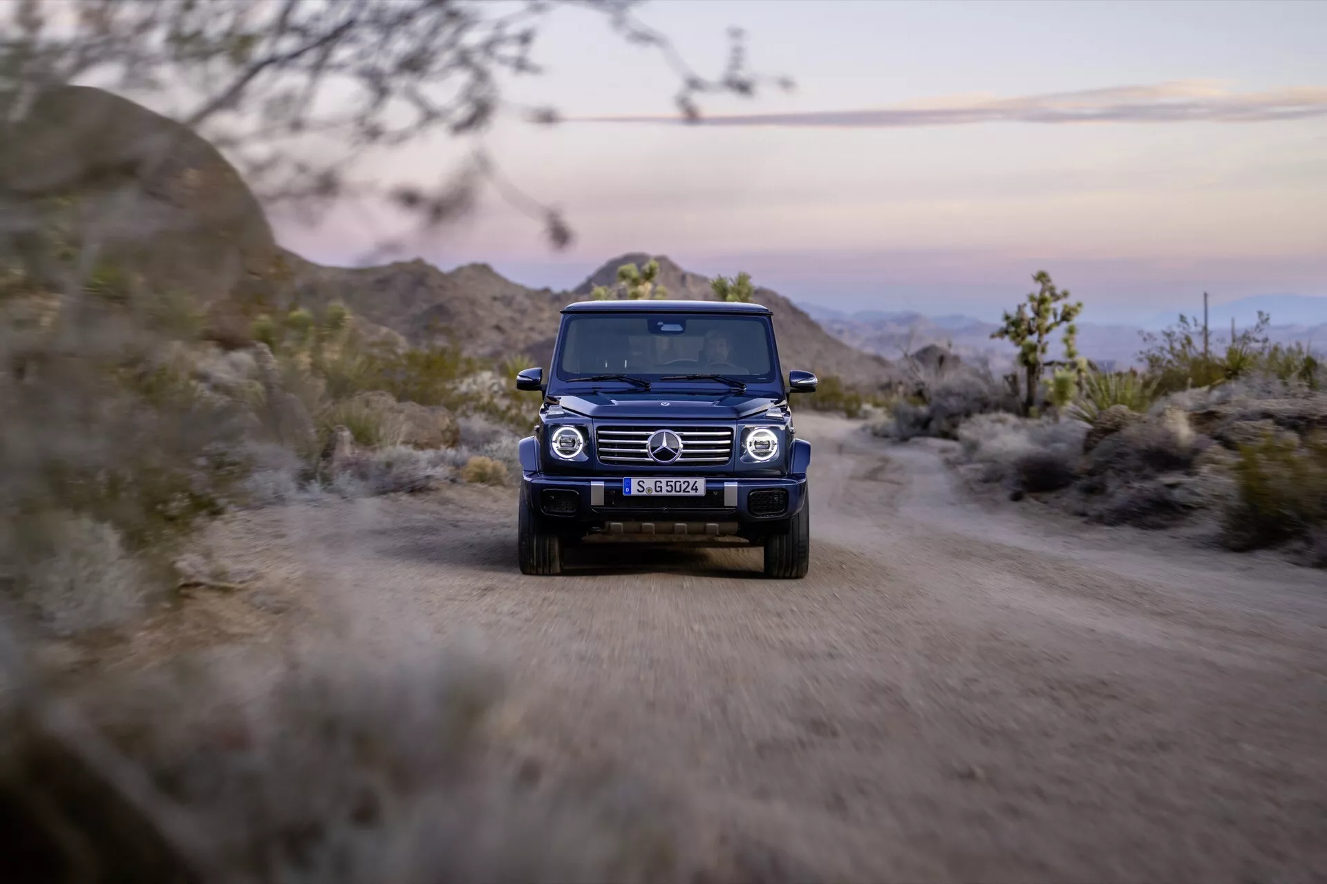 SUV Mercedes-Benz Classe G 2024 roulant sur un chemin de terre avec les phares allumés, soulignant son design robuste et ses capacités tout-terrain dans une vue de face au crépuscule