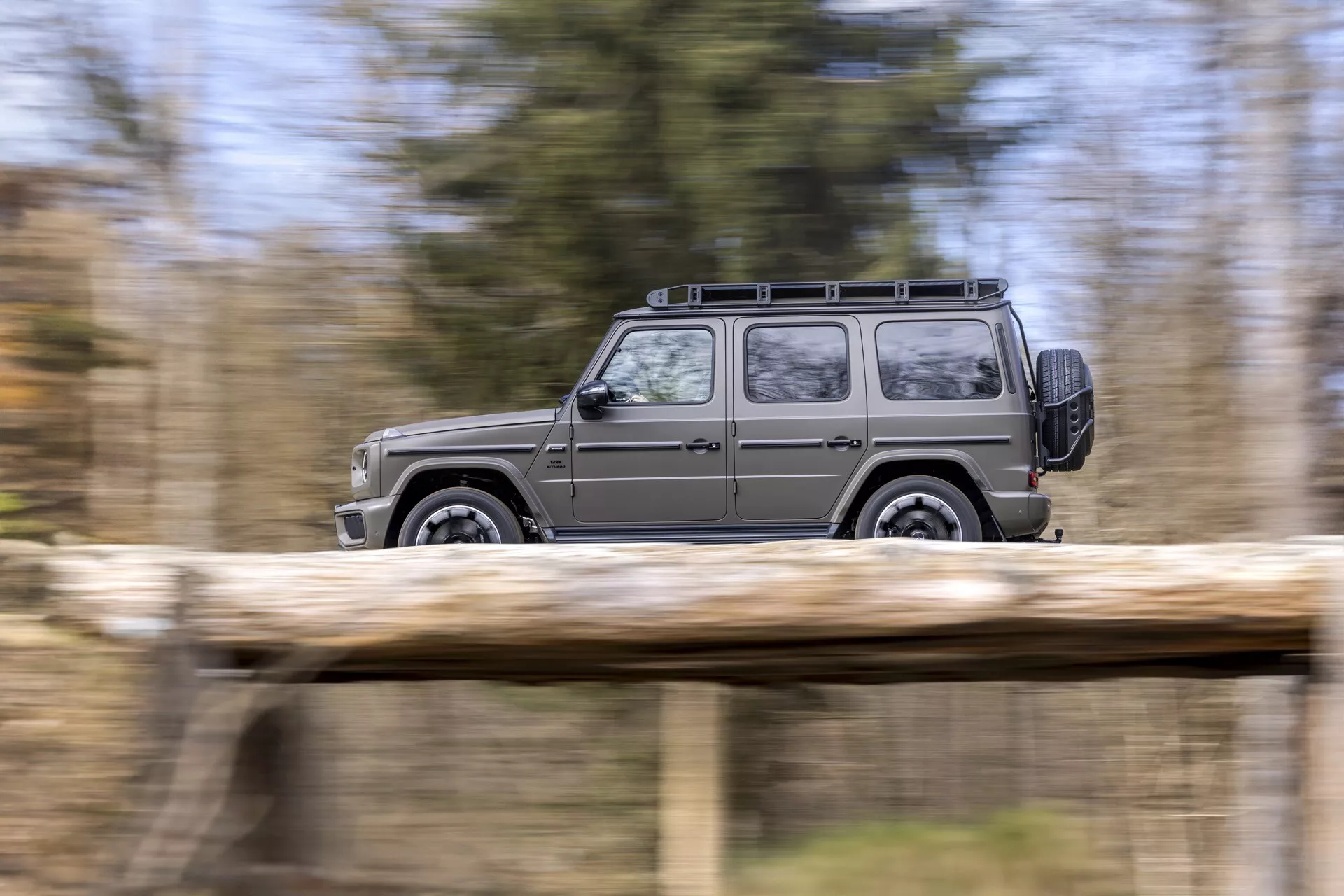 Vue latérale dynamique du SUV Mercedes-Benz Classe G (G-Klasse) 2024 montrant son mouvement avec un arrière-plan naturel flou, mettant en avant son design carré, porte-bagages et roues en alliage