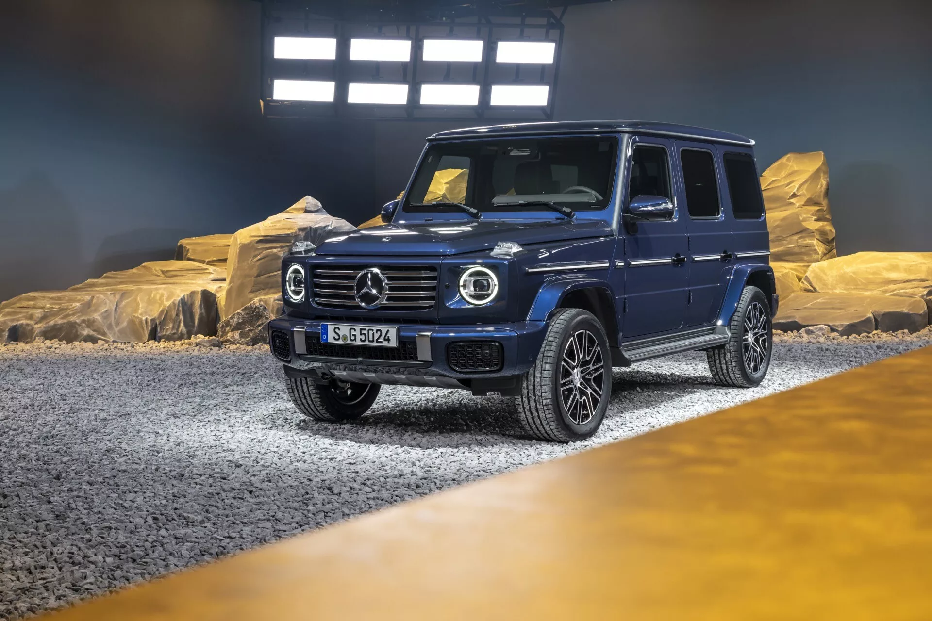 SUV de luxe Mercedes-Benz Classe G (G-Klasse) 2024 dans un showroom avec éclairage dramatique, offrant une vue diagonale de l'avant et du côté soulignant ses phares LED ronds, calandre proéminente avec grand emblème, pare-chocs avant robuste avec technologie avancée, jantes en alliage à multiples branches, et un design robuste mais sophistiqué adapté pour une utilisation tout-terrain et urbaine, immatriculé 'S-GO524'.