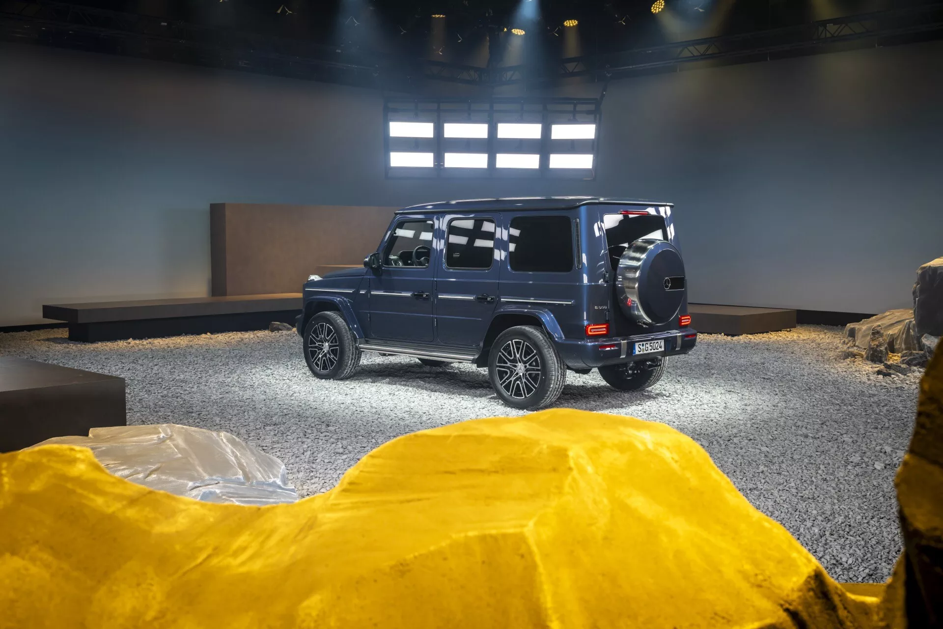SUV Mercedes-Benz Classe G (G-Klasse) 2024 dans un showroom, vue arrière et latérale mettant en avant les capacités tout-terrain avec une roue de secours montée à l'arrière, des jantes en alliage et une garde au sol élevée sur un fond mis en scène