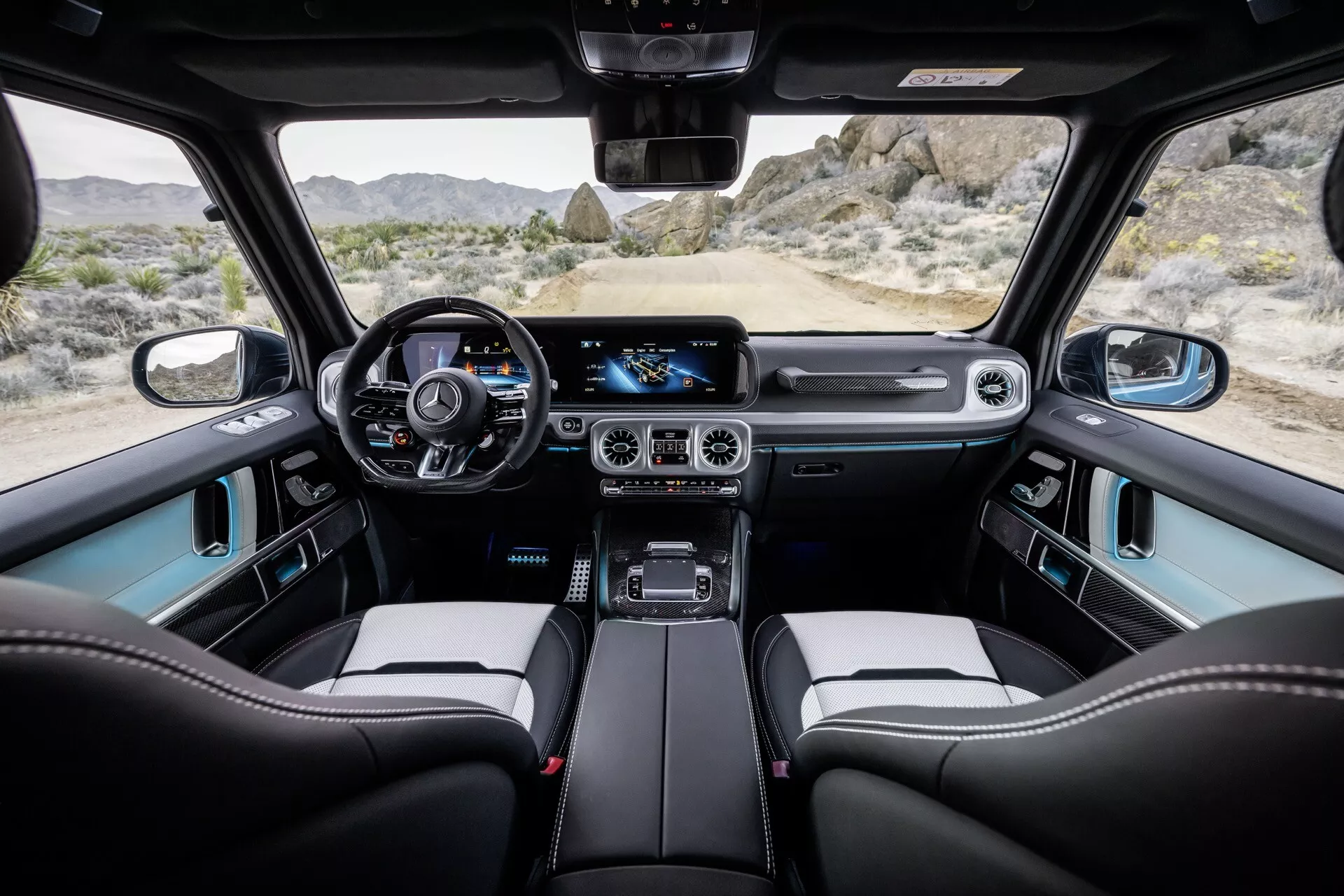 Vue intérieure de la Mercedes-Benz Classe G 2024 depuis le siège arrière, montrant le tableau de bord, le cluster d'instruments numérique, le volant en cuir avec commandes, le grand système d'infodivertissement tactile, le contrôle de climatisation bi-zone, le levier de vitesse robuste, les sièges avant premium baquets et le toit panoramique.
