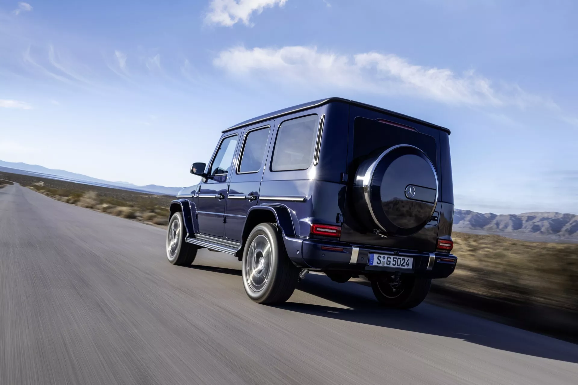 SUV de luxe Mercedes-Benz G-Klasse 2024 de couleur sombre roulant sur une route asphaltée dans un environnement semblable à celui d'un désert, présentant sa vue de quart arrière avec jante brillante, design cubique et caractéristiques tout-terrain comme pneu de secours et feux arrière LED.