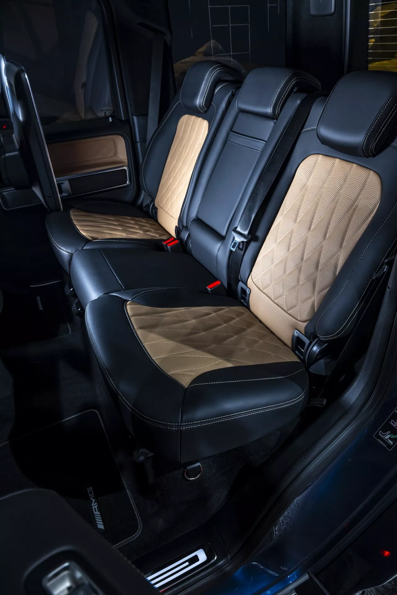 Compartiment passager arrière du Mercedes-Benz G-Klasse (G 465) 2024 présentant des sièges haut de gamme noirs et beige clair avec motifs losangés, clips de ceinture de sécurité rouges, panneaux de porte avec finitions en bois et fonctionnalités avancées de la cabine, mettant en avant la conception intérieure de luxe et le confort.