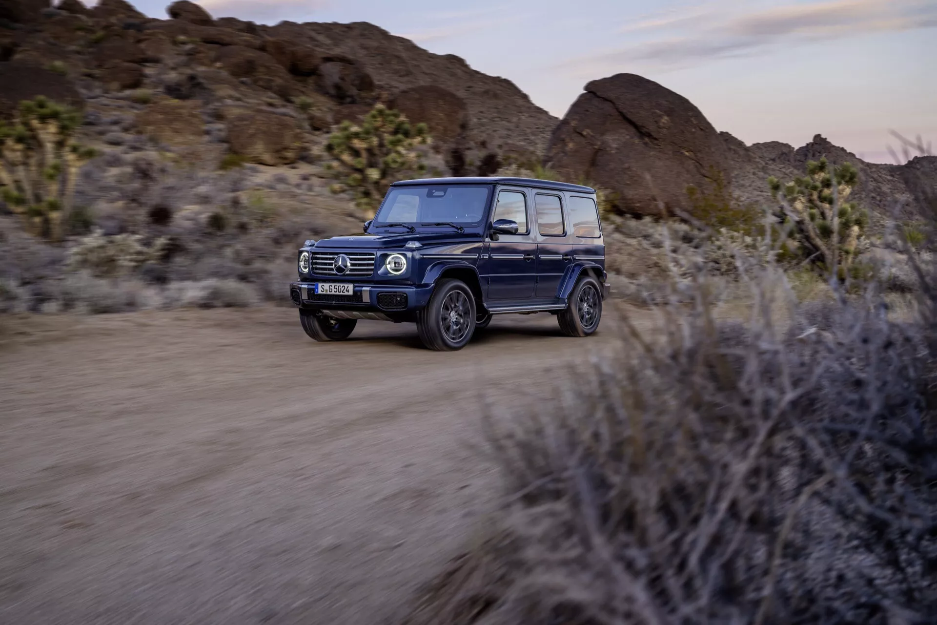 SUV Mercedes-Benz Classe G 2024 roulant sur une route non pavée dans le désert, doté de phares LED, de technologie adaptative, d'un intérieur luxueux, d'un design tout-terrain et de fonctions de sécurité avancées, avec un flou de mouvement pour souligner les capacités dynamiques