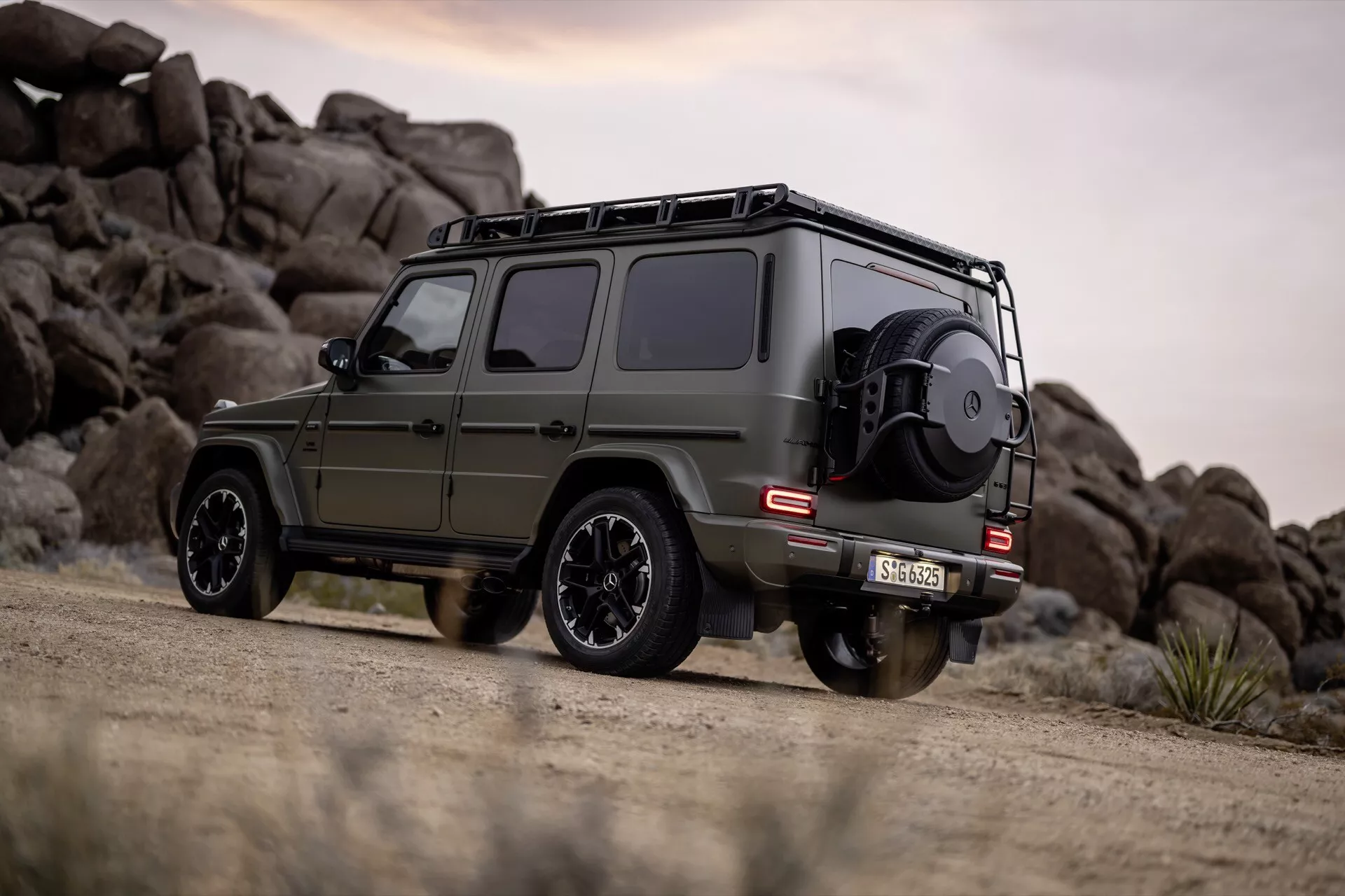 Mercedes-Benz Classe G (G-Klasse 465) 2024 en vert olive mat, présentant sa vue latérale et arrière avec des caractéristiques tout-terrain, stationnée sur un chemin de terre devant un paysage rocheux en arrière-plan.