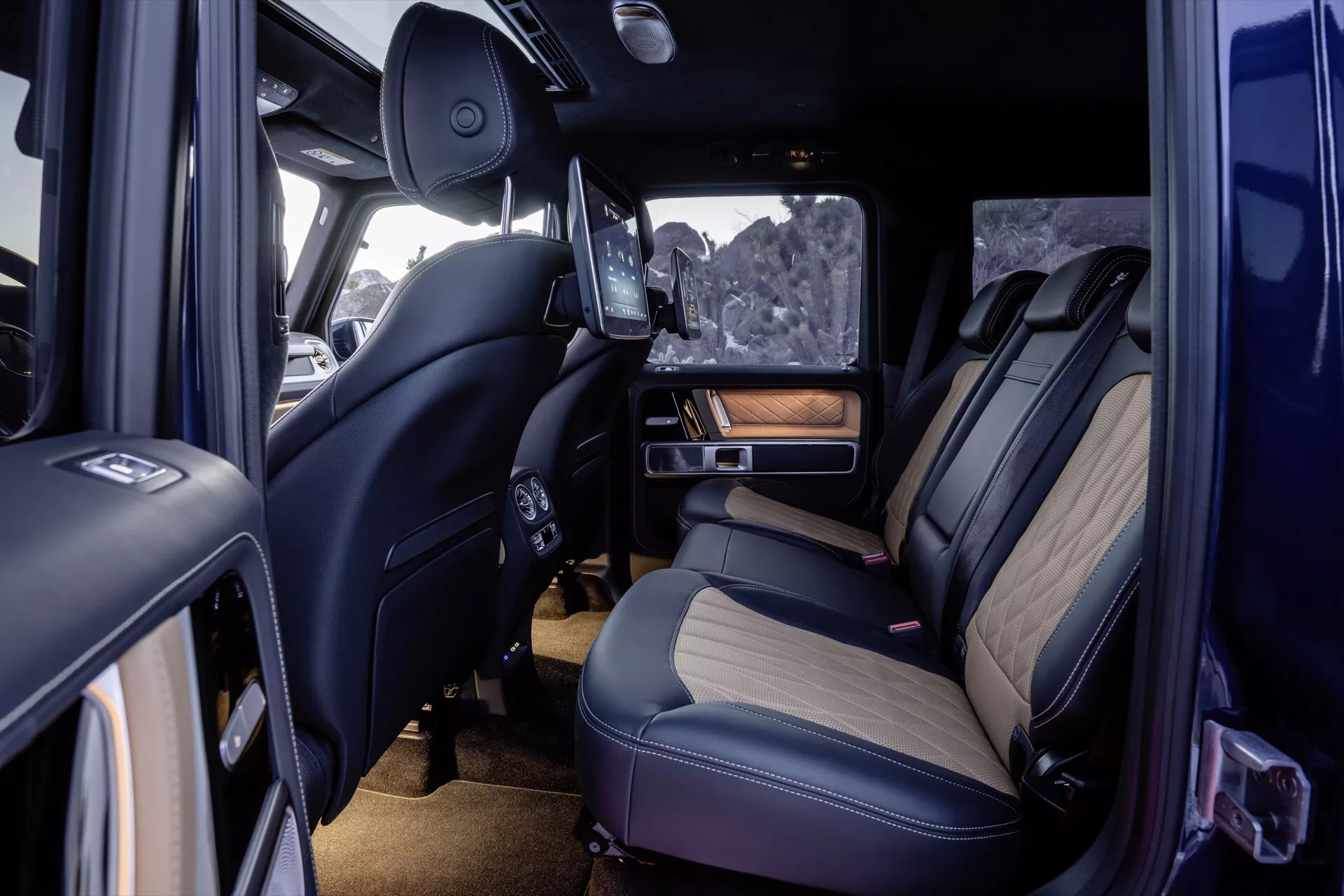 Vue intérieure du Mercedes-Benz Classe G 465 2024 montrant les sièges arrière premium, coutures détaillées, matériaux de luxe avec des accents de bois, fonctionnalités technologiques avancées incluant un écran de divertissement rétractable, éclairage d'ambiance et contrôle climatique depuis une perspective de porte latérale passager ouverte