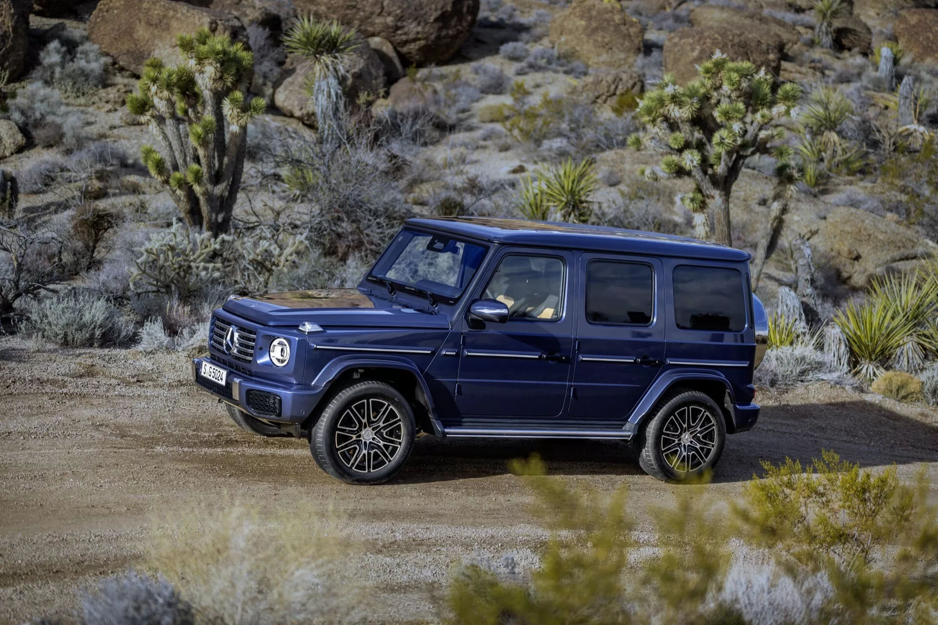 SUV sombre Mercedes-Benz Classe G 465 de 2024, garé hors-route sur un chemin non pavé entouré de végétation sèche, présentant son design robuste et carré, grandes roues à multiples rayons et barres de toit adaptées pour l'aventure et les capacités tout-terrain de luxe.