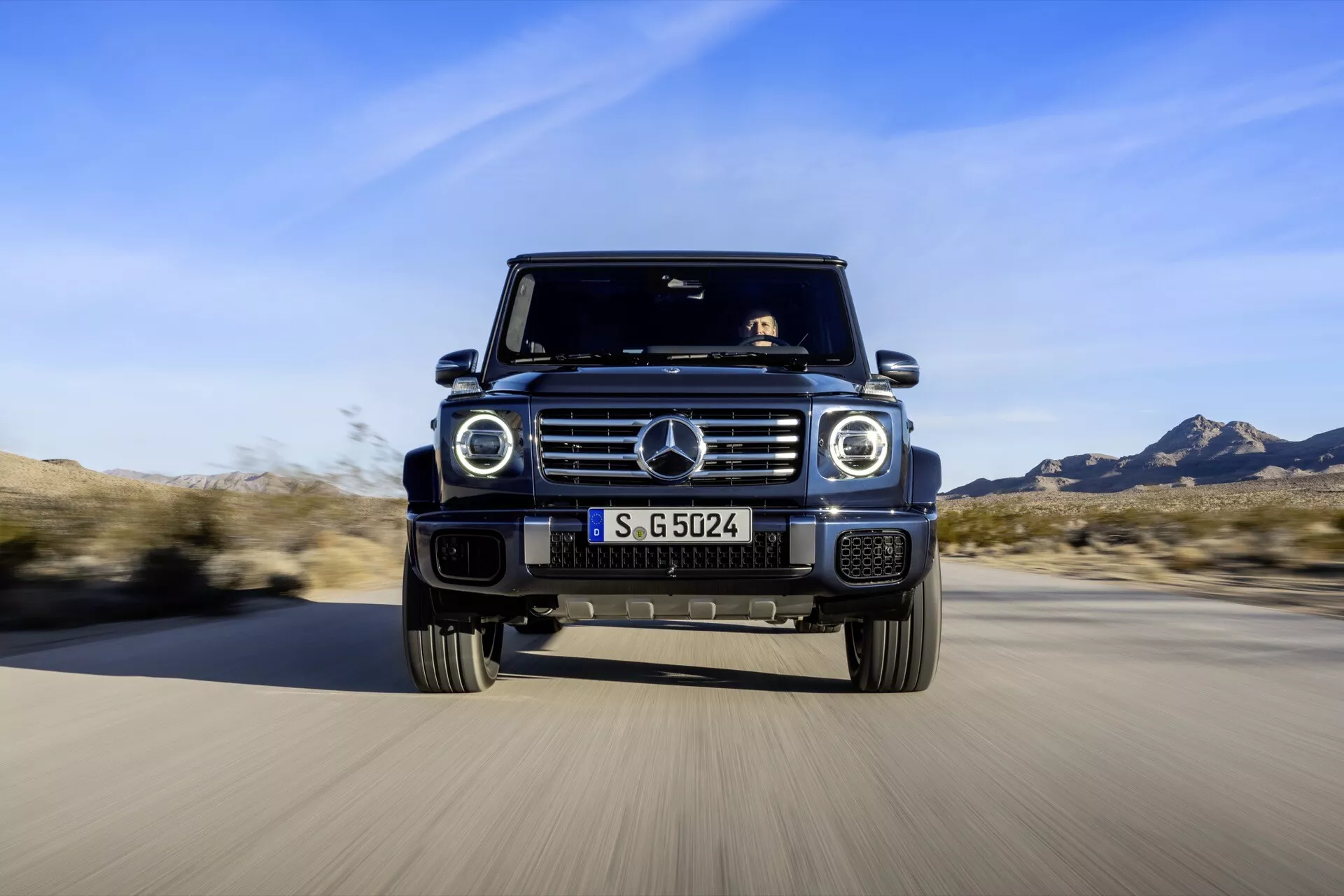 SUV Mercedes-Benz Classe G 2024 s'approchant sur une route, montrant un angle bas de sa grille emblématique, des lumières LED circulaires, une plaque de protection sur le pare-chocs avant, une caméra frontale et sa silhouette carrée distinctive sur un fond flou, soulignant la présence dynamique et les caractéristiques avancées du véhicule.