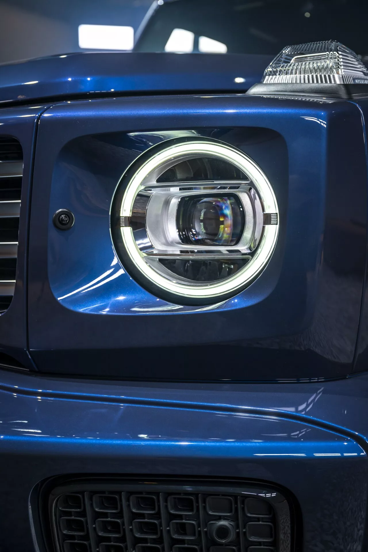 Gros plan sur l'ensemble phare gauche de la Mercedes-Benz G-Klasse 2024, présentant un design moderne de projecteur LED avec des accents chromés sur une carrosserie bleue métallisée