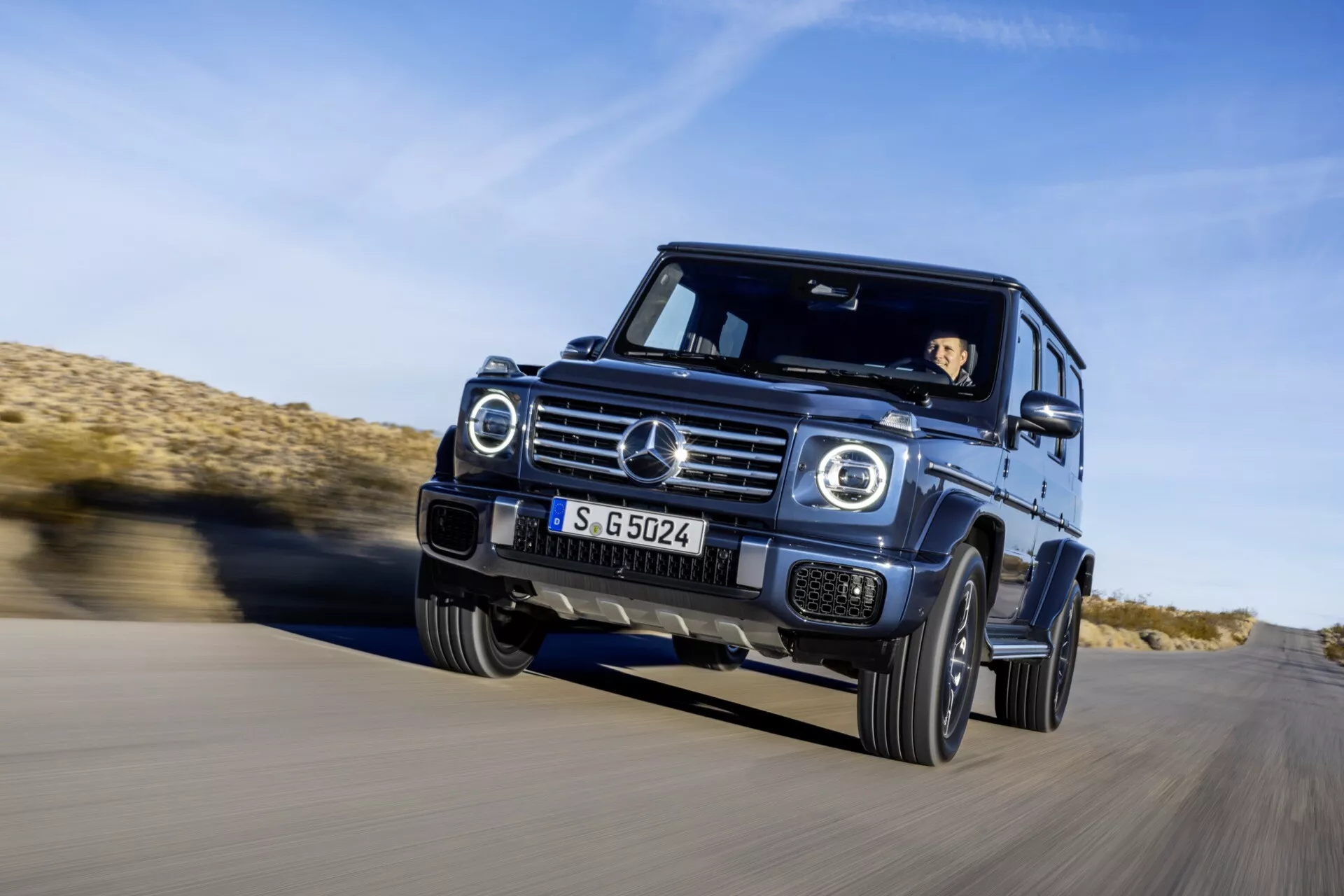 SUV Mercedes-Benz Classe G 2024 roulant sur asphalte dans un environnement désertique, vue avant trois quarts mettant en évidence le mouvement dynamique, phares LED, grille chromée et capacités tout-terrain
