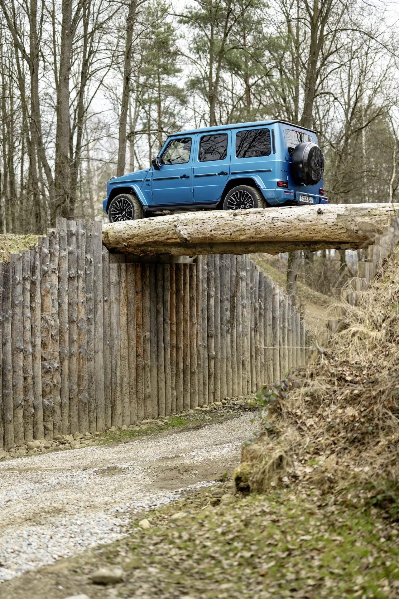 SUV bleu Mercedes-Benz G-Klasse 2024 sur plateforme en bois surélevée en milieu tout-terrain, montrant les vues arrière et latérale avec roue de secours visible