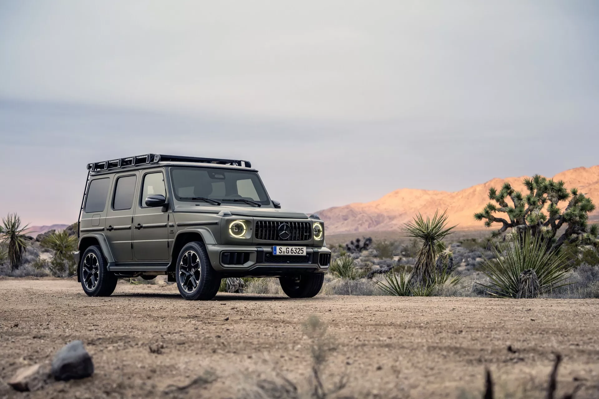 SUV Mercedes-Benz G-Klasse 2024 dans un cadre extérieur accidenté, montrant un angle avant trois quarts avec vue détaillée de la calandre, des phares et des roues tout-terrain sur un chemin de terre