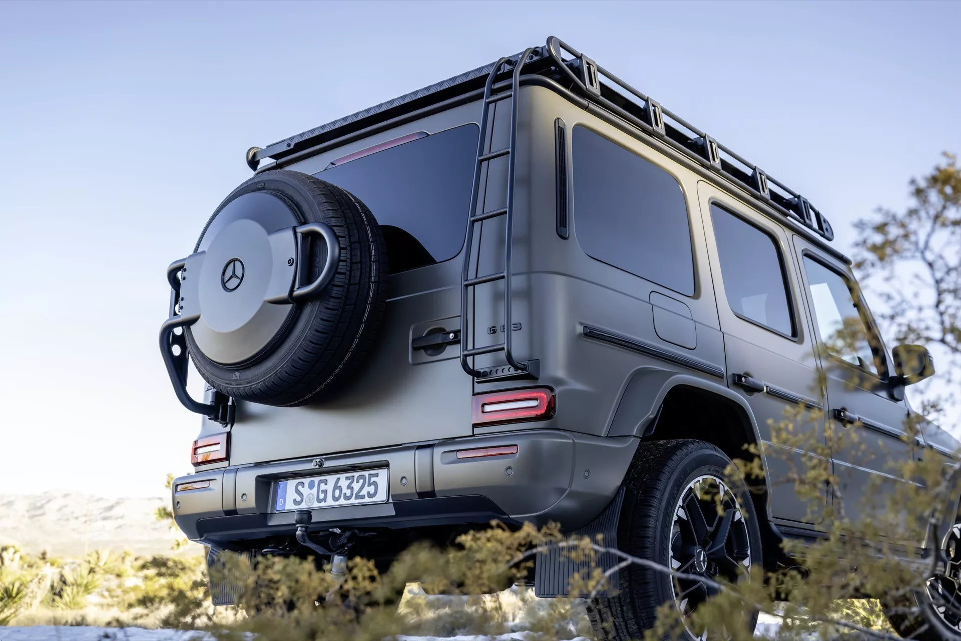 Vue arrière trois quarts du SUV Mercedes-Benz G-Klasse 2024 montrant le côté droit, prise sous un angle bas pour souligner sa posture puissante avec des détails centrés incluant des feux arrière LED, un pneu arrière monté avec couverture à emblème, un design robuste indiquant une capacité tout-terrain, des capteurs de stationnement, un crochet de remorquage, et un porte-bagages sur une finition métallique sous un éclairage ambiant.