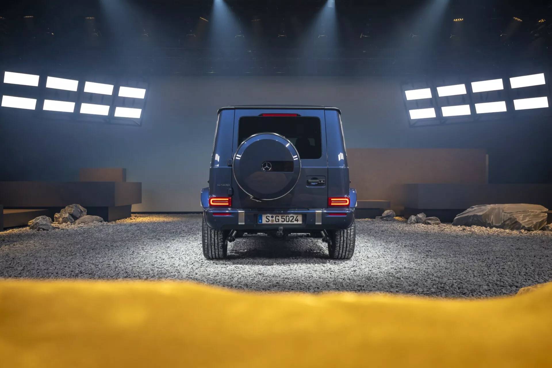 Vue arrière du SUV Mercedes-Benz Classe G (G-Klasse) 2024 dans un décor de studio simulant un environnement tout-terrain, mettant en avant la hauteur, les feux arrière, la roue de secours et l'éclairage stratégique