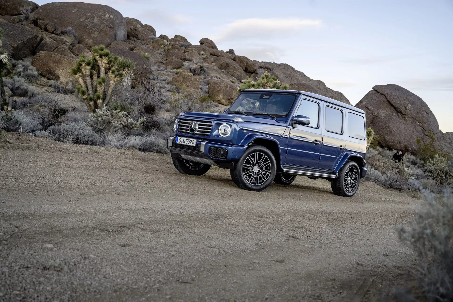 SUV bleu Mercedes-Benz Classe G 465 de 2024 garé sur un terrain accidenté, affichant un design carré, une garde au sol élevée et exposant la capacité tout-terrain du véhicule depuis une vue de trois quarts avant qui met en lumière les jantes en alliage et les pneus tout-terrain