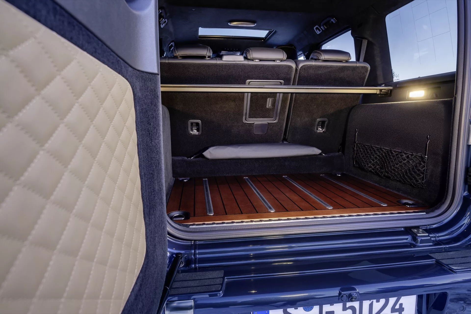 Vue rapprochée de l'espace de coffre intérieur de la Mercedes-Benz G-Klasse 2024, mettant en lumière son revêtement de moquette de luxe, ses lattes de sol en bois, ses crochets de chargement en métal, et sa poche de rangement en filet, à côté des sièges arrière de luxe et de l'éclairage intégré du coffre