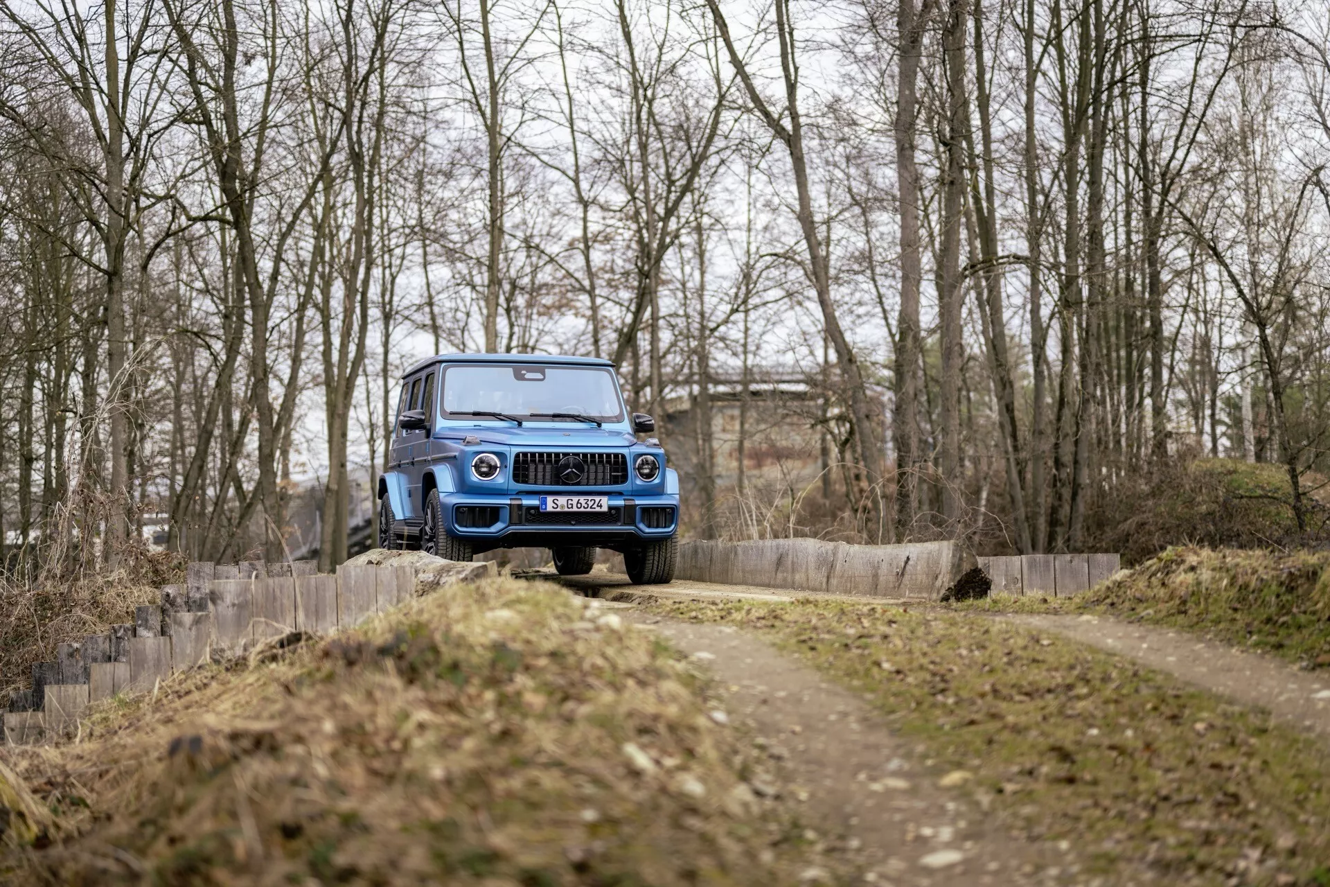 Véhicule tout-terrain bleu Mercedes-Benz G-Klasse 2024 en mouvement sur un chemin de terre, vue de trois quarts avant présentant la calandre avant, le capot, le pare-brise et la garde au sol élevée, dans un cadre forestier sans neige