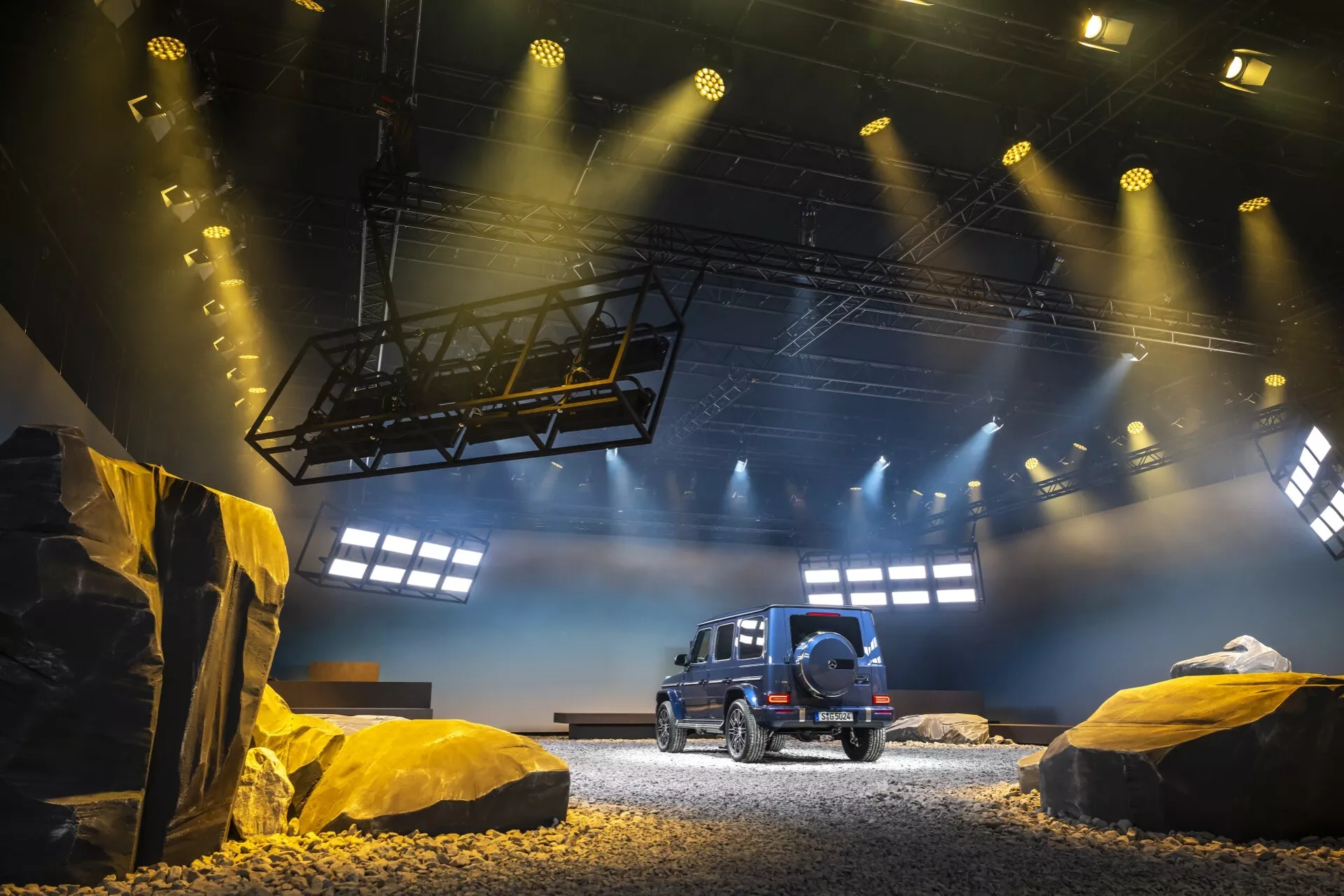 SUV Mercedes-Benz G-Klasse 2024 présenté dans un cadre tout-terrain simulé, montrant la vue arrière et de côté du véhicule avec profil latéral détaillé, lunette arrière, feux arrière LED et une roue de secours montée sur le hayon, sous un éclairage dramatique d'exposition