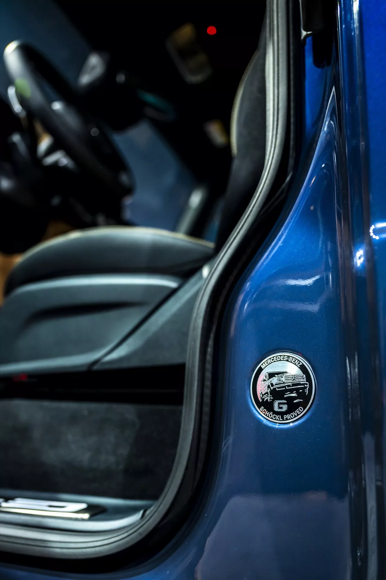 Image en gros plan montrant le badge sur le montant B de la Mercedes-Benz G-Klasse 2024, avec un extérieur bleu vif et un design intérieur sportif noir, mettant en lumière les attributs d'un véhicule haut de gamme axé sur la marque et les matériaux de haute qualité.