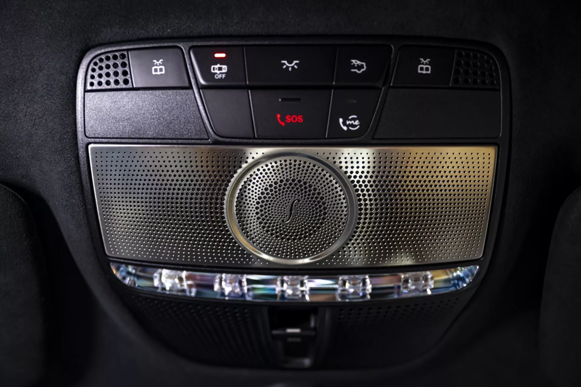Vue rapprochée de la console supérieure de la Mercedes-Benz G-Klasse 2024 (465) avec boutons de commande, haut-parleur central, options d'éclairage intérieur, services d'urgence et fonctions personnalisables, soulignant le système multimédia avancé et les détails de conception sophistiqués