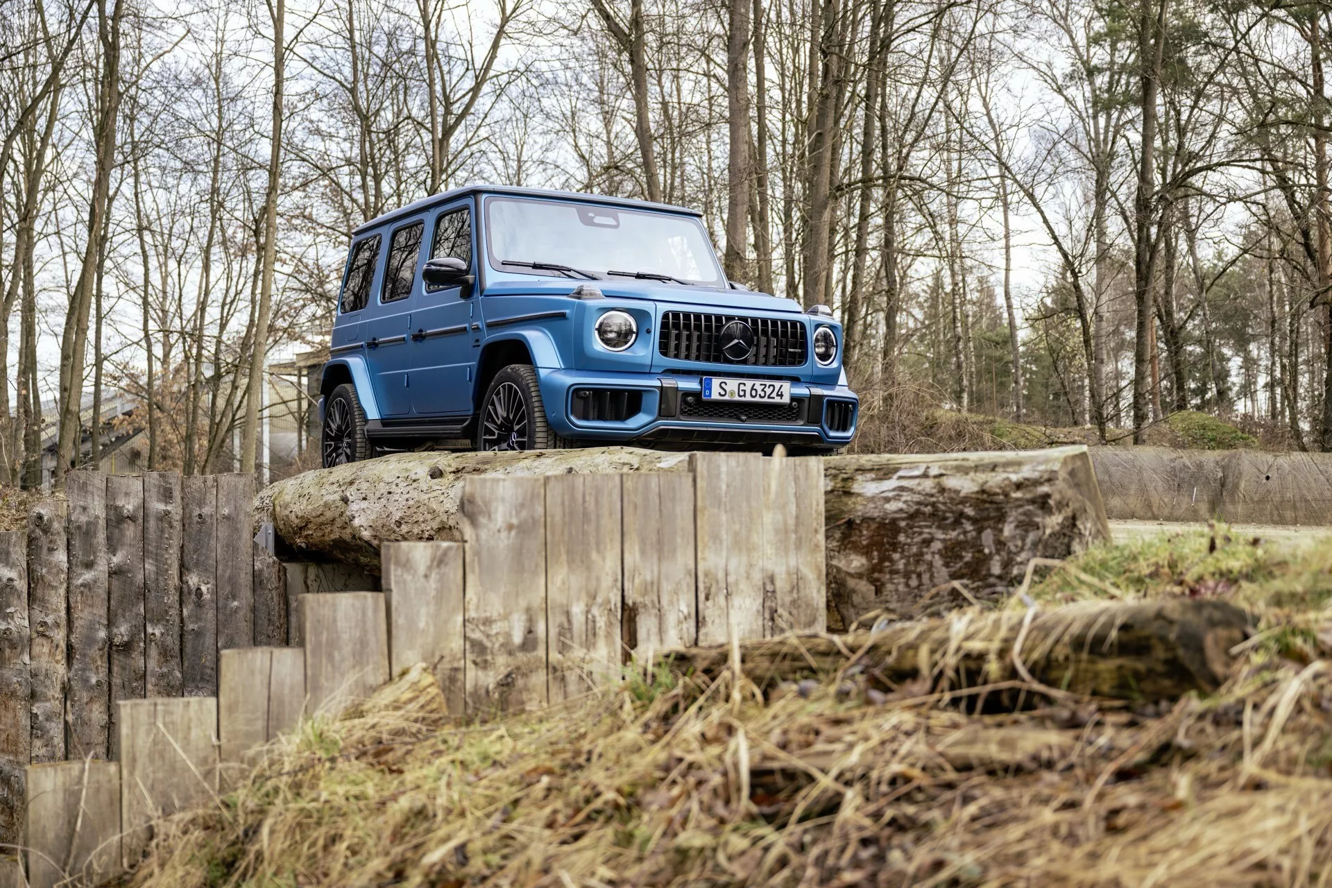 SUV Mercedes-Benz G-Klasse (Classe G) 465 de 2024 démontrant ses capacités tout-terrain sur une rampe en bois à plusieurs niveaux dans un terrain accidenté, mettant en évidence sa grande garde au sol, sa grille avant robuste, ses phares LED et ses pneus à profondes sculptures adaptés pour une utilisation en terrains difficiles