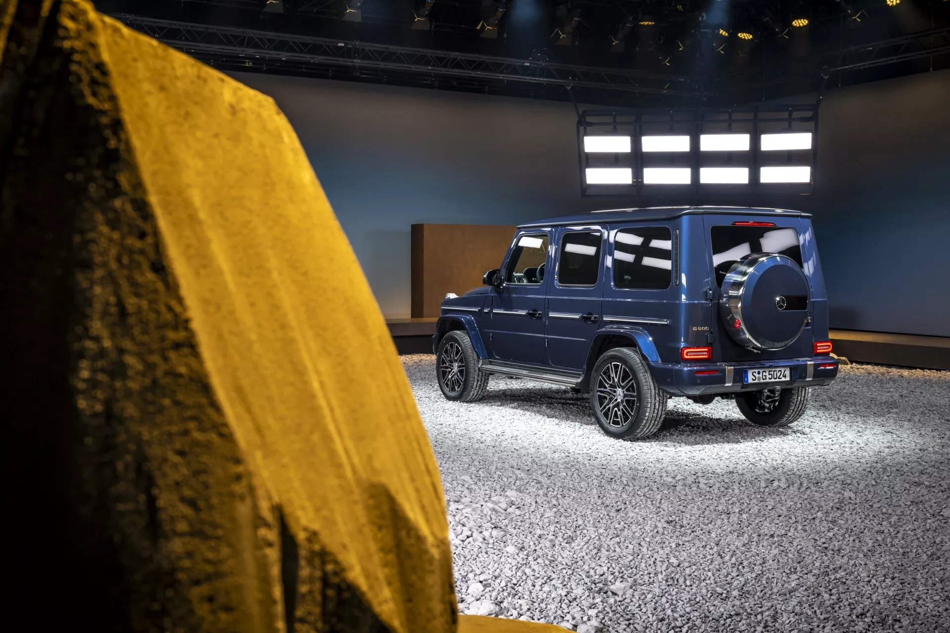 Vue de côté arrière de la Mercedes-Benz Classe G (G 465) 2024 dans un cadre de studio, mettant en avant son design robuste et luxueux, ses feux arrière à LED et sa roue de secours sur un bleu métallique profond