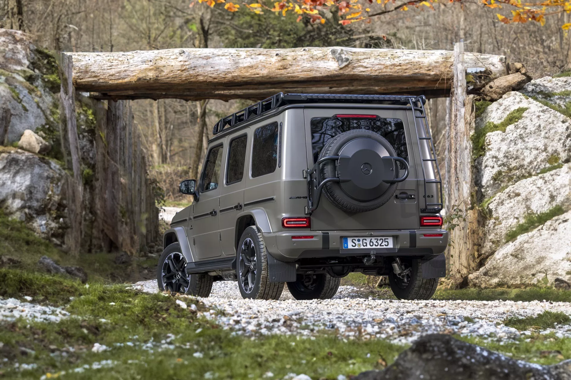 Vue arrière en angle du SUV Mercedes-Benz G-Klasse 465 2024, soulignant la hauteur et la garde au sol sur terrain rocheux, équipé d'un pneu de secours, d'un récepteur d'attelage de remorque et démontrant ses capacités en tout-terrain