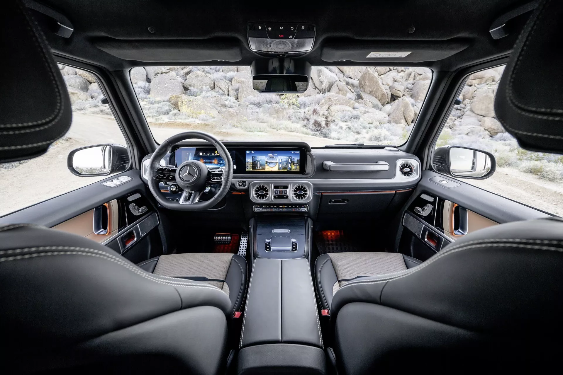 Vue intérieure de la Mercedes-Benz Classe G (G-Klasse 465) 2024 depuis le siège arrière montrant des sièges en cuir noir, volant moderne avec commandes, tableau de bord en cuir et métal, cluster d'instruments numérique, console centrale avec finitions métalliques, grand écran tactile d'infodivertissement, boutons de contrôle climatique avec finition métallique, commandes d'éclairage ambiant au plafond, système de haut-parleurs haut de gamme, toit ouvrant panoramique, rétroviseur central, et larges fenêtres améliorant la visibilité et la sensation de luxe.