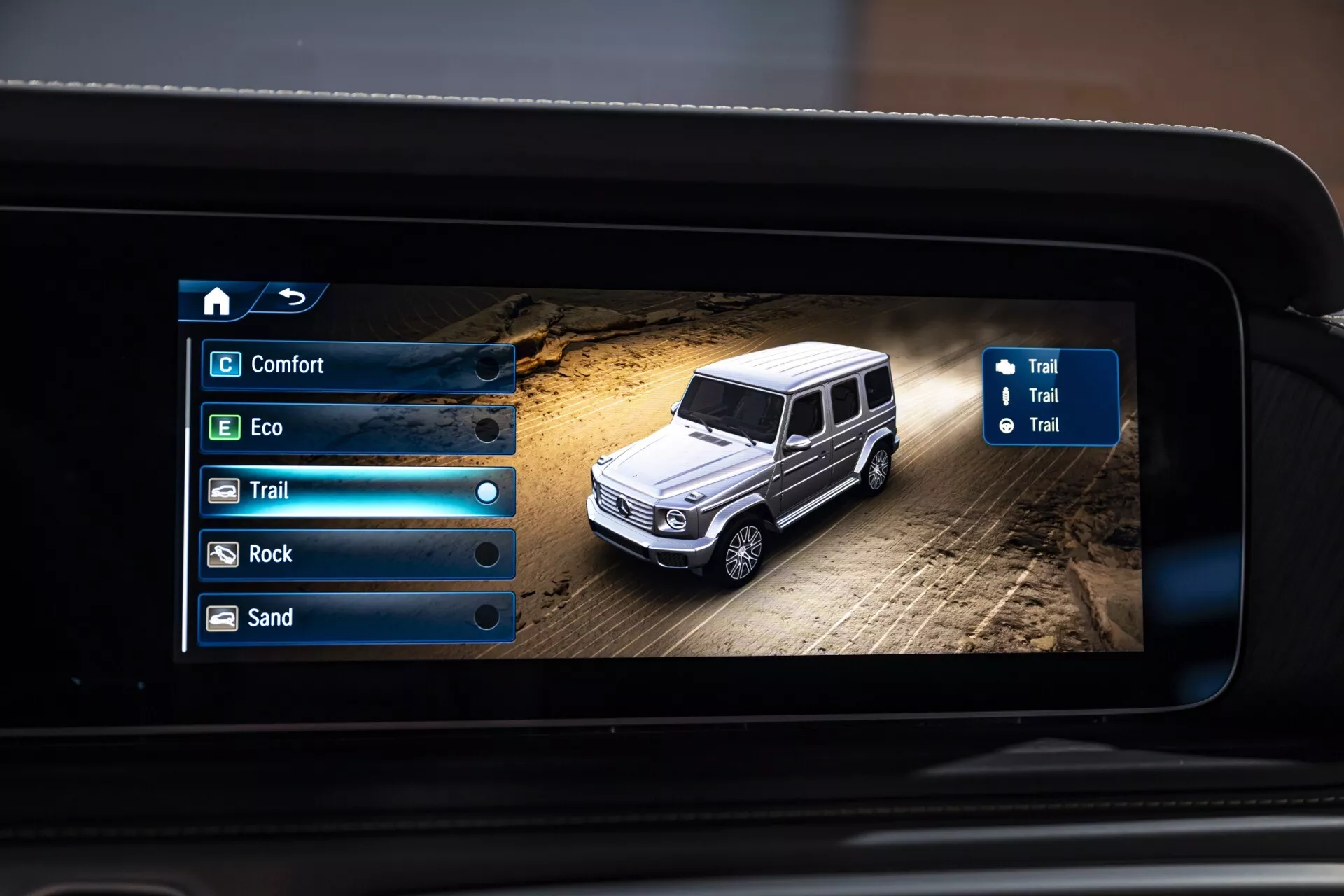 Affichage numérique de Mercedes-Benz Classe G 2024 en configuration tout-terrain montrant les modes de conduite sélectionnables sur l'écran du système d'infodivertissement, vue de dessus d'un SUV de luxe sur un terrain poussiéreux avec navigation et options de mode