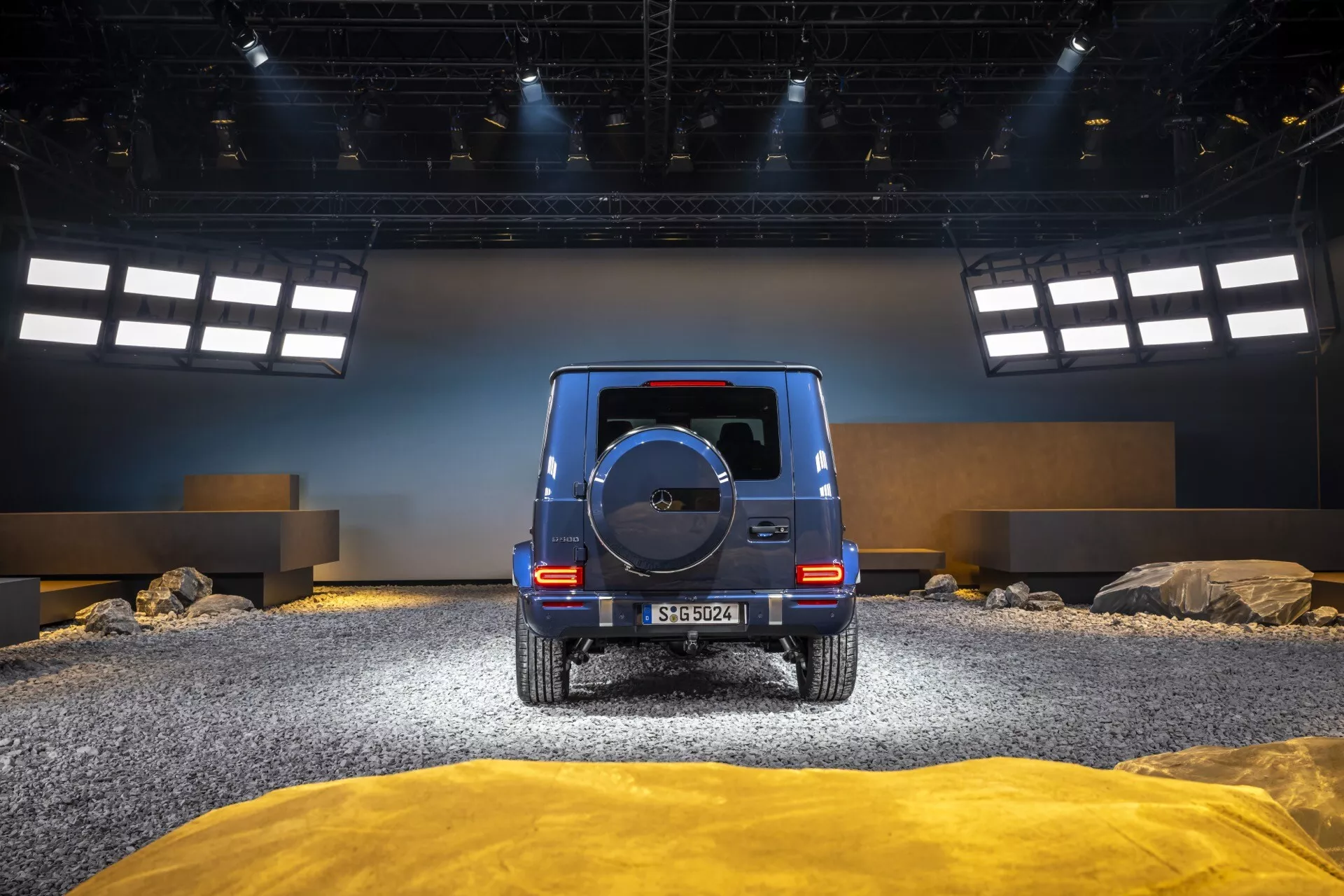 Vue arrière du Mercedes-Benz Classe G (G 465) 2024 dans un décor tout-terrain avec roches et sol texturé, mettant en avant des feux arrière LED, une roue de secours et une finition brillante.