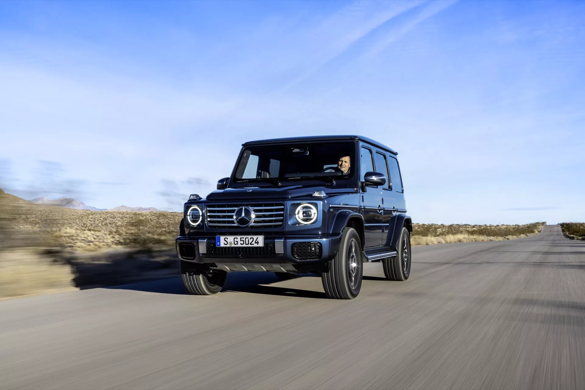 SUV de luxe Mercedes-Benz Classe G 2024 en mouvement, capturée depuis un angle frontal trois quarts montrant les phares LED, les feux de jour circulaires, le design robuste et les caractéristiques technologiques avancées contre un arrière-plan de route désertique