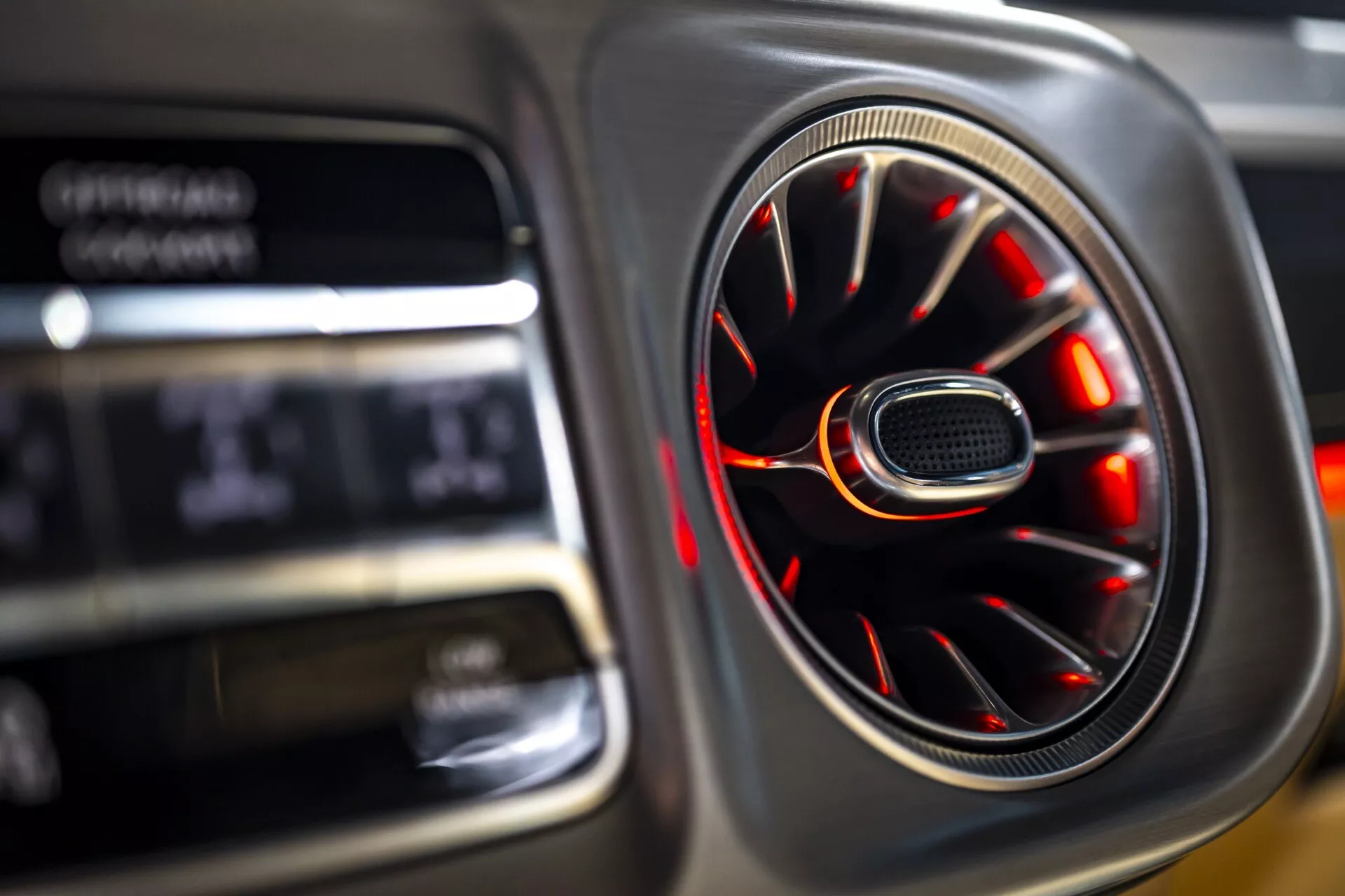 Vue rapprochée de l'intérieur d'une Mercedes-Benz G-Klasse 2024 (465) montrant une sortie de ventilation finition chrome avec éclairage d'ambiance rouge et arrière-plan flou de l'affichage numérique soulignant le luxe et le design