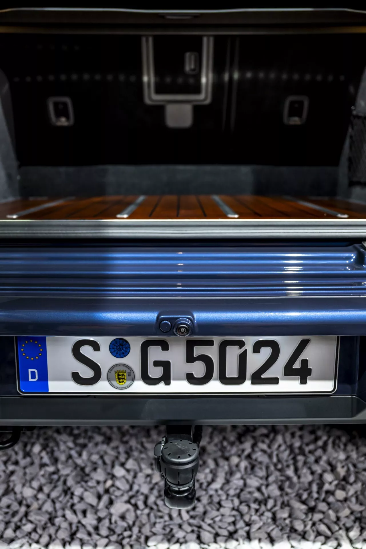 Gros plan détaillé sur la section arrière du Mercedes-Benz G-Klasse 2024 mettant en évidence la zone du coffre, le revêtement du coffre en bois de luxe, la caméra de recul intégrée au-dessus de la plaque d'immatriculation de style européen avec le 'D' de l'Allemagne, et le crochet de remorquage en dessous, le tout capturé d'un angle bas et orienté vers le haut soulignant la peinture métallisée premium et l'esthétique de conception du véhicule.