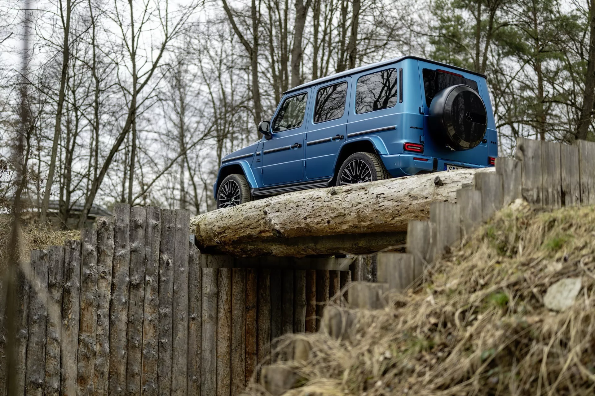 Mercedes-Benz Classe G 2024 en bleu, vue de côté mettant en avant son design carré, sa suspension tout-terrain et sa garde au sol sur une surface surélevée, avec un focus sur ses caractéristiques de durabilité et de luxe sur fond d'arbres nus et d'une clôture en bois